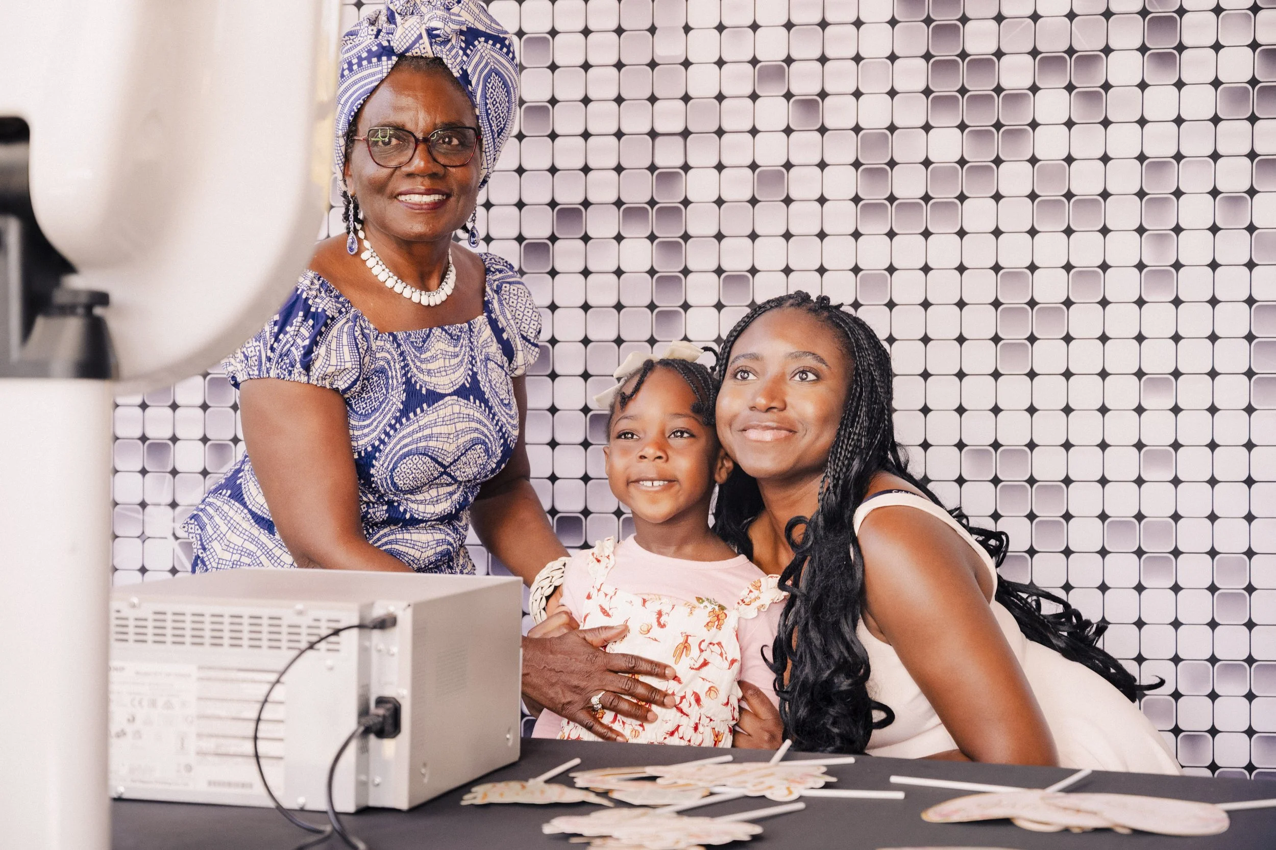 A woman with glasses and a patterned headwrap standing behind two young girls, all smiling for the camera in front of a black and white geometric tiled wall.
