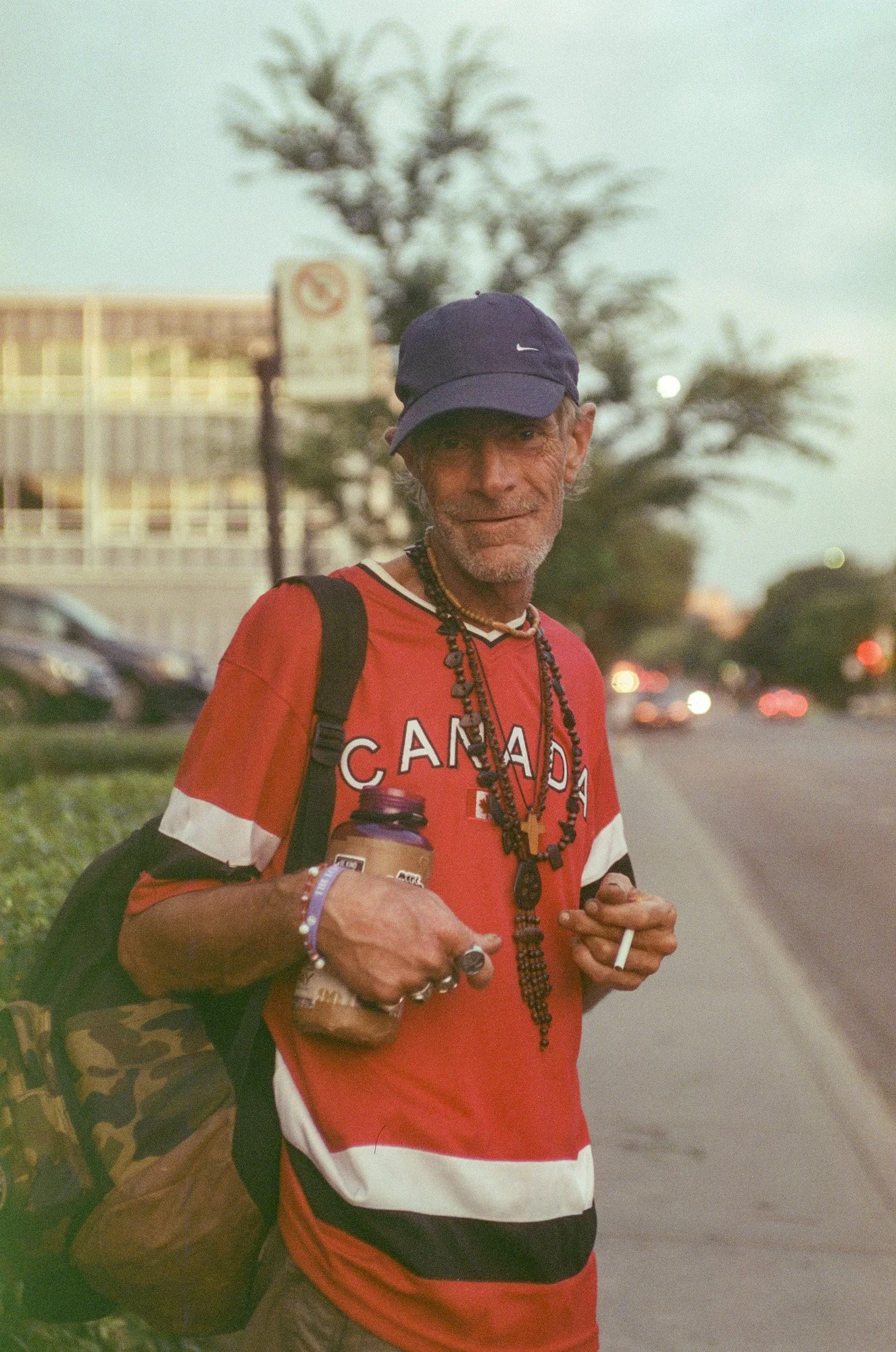 An older man wearing a red Canada football jersey, a dark blue Nike baseball cap, and multiple necklaces, carrying a camo backpack and holding a drink and a cigarette, standing outdoors during sunset with a blurred background of cars and trees.