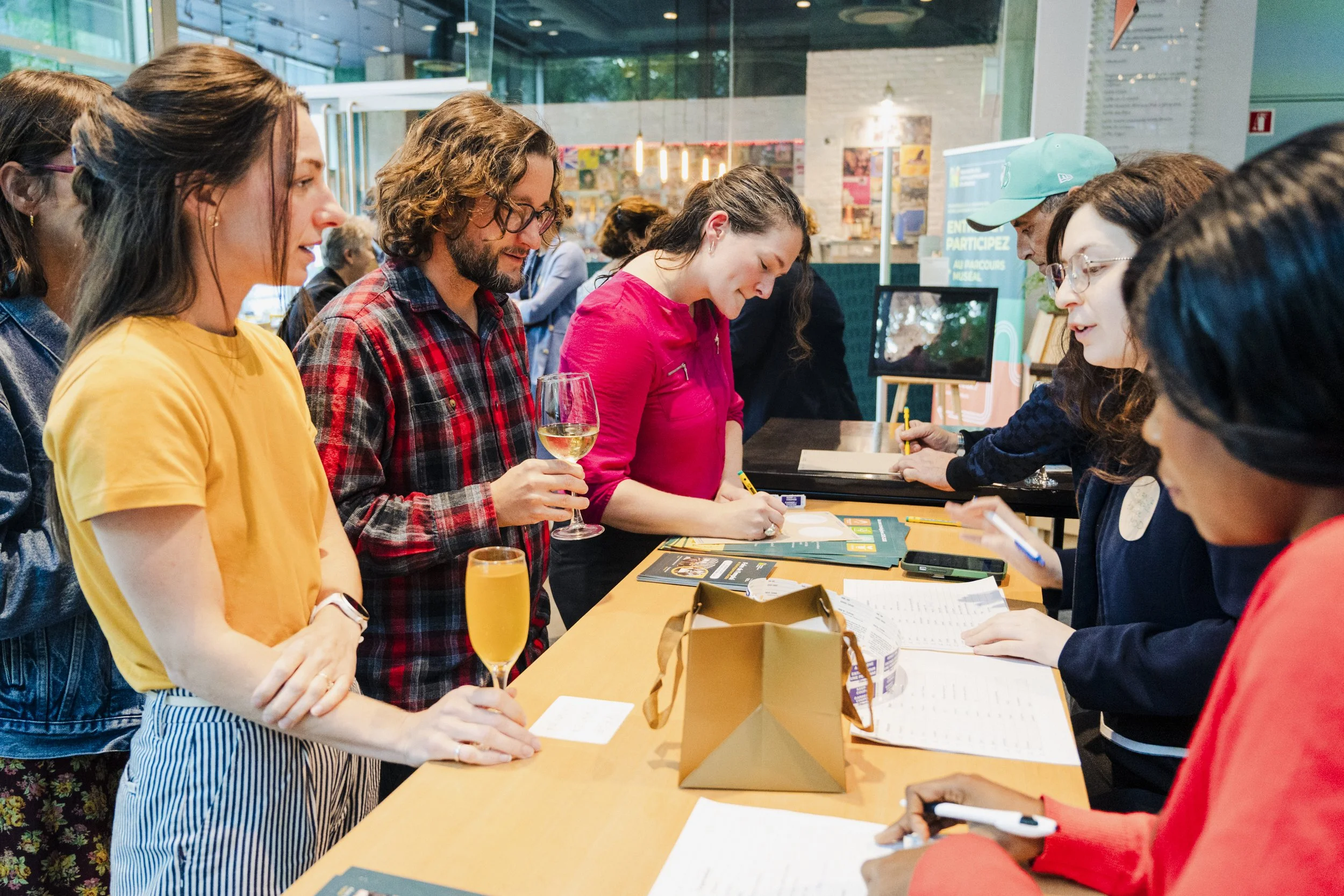 A group of people gathered around a counter at an indoor event, with some signing or filling out forms, and others observing. One woman has a glass of white wine in hand. There are brochures, papers, and bags on the counter, with a brightly lit, mode
