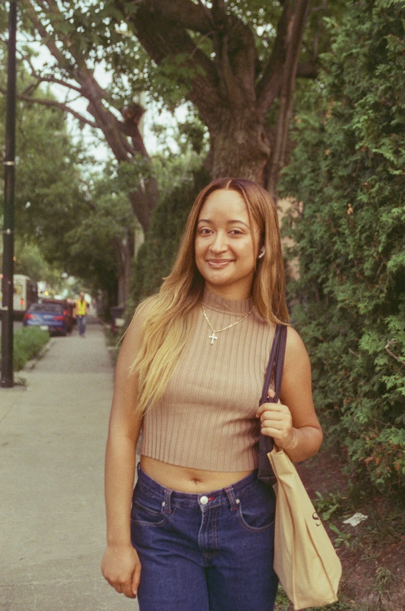 A young woman smiling and walking on the sidewalk next to a green bush and large tree, holding a beige tote bag, wearing a beige sleeveless crop top, blue jeans, and AirPods, with a street and parked cars in the background.