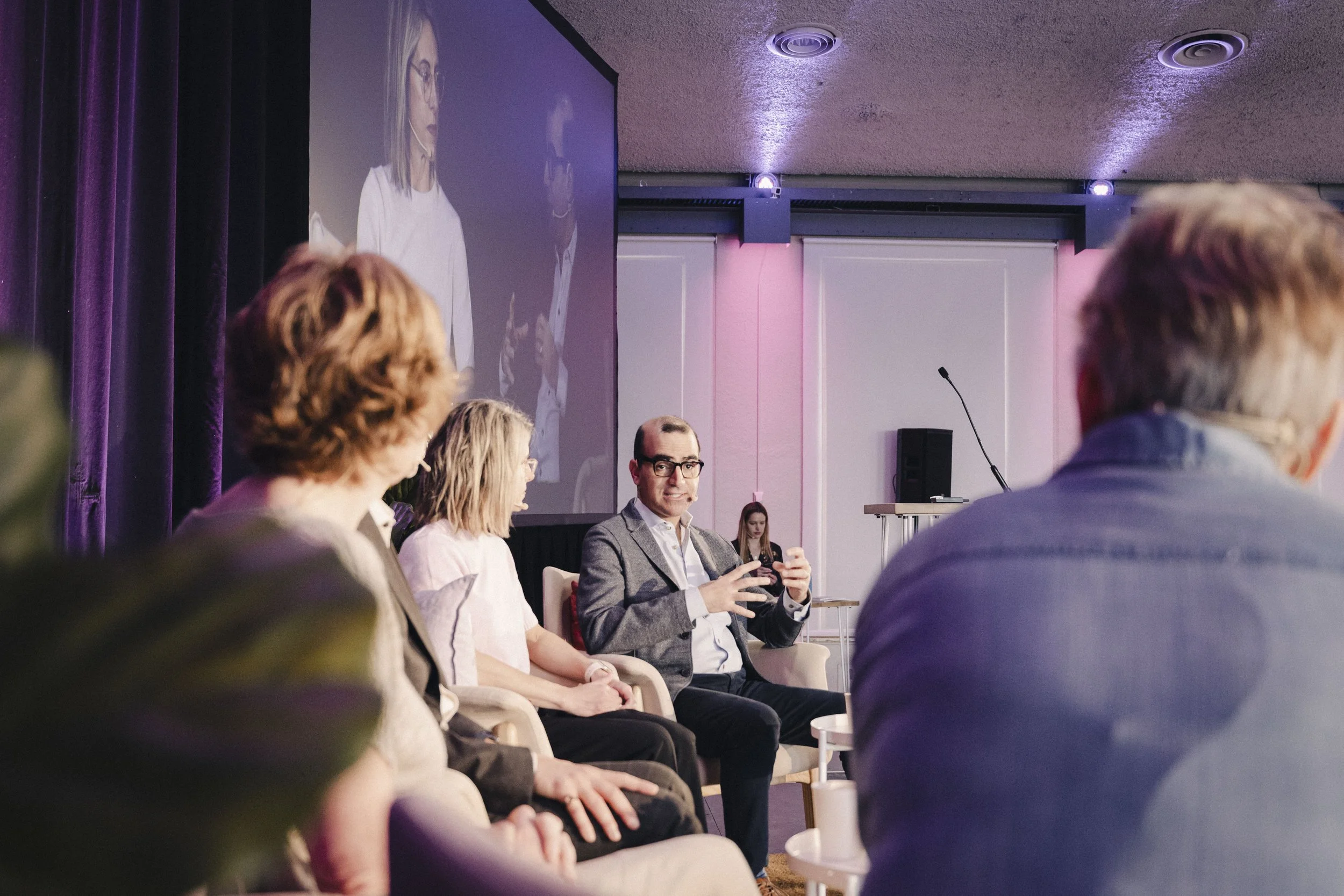A panel discussion at an indoor conference with five people sitting on stage, including a man in a gray suit speaking into a microphone. A large screen behind them displays the image of the woman and the man on stage, with purple curtains and pink li