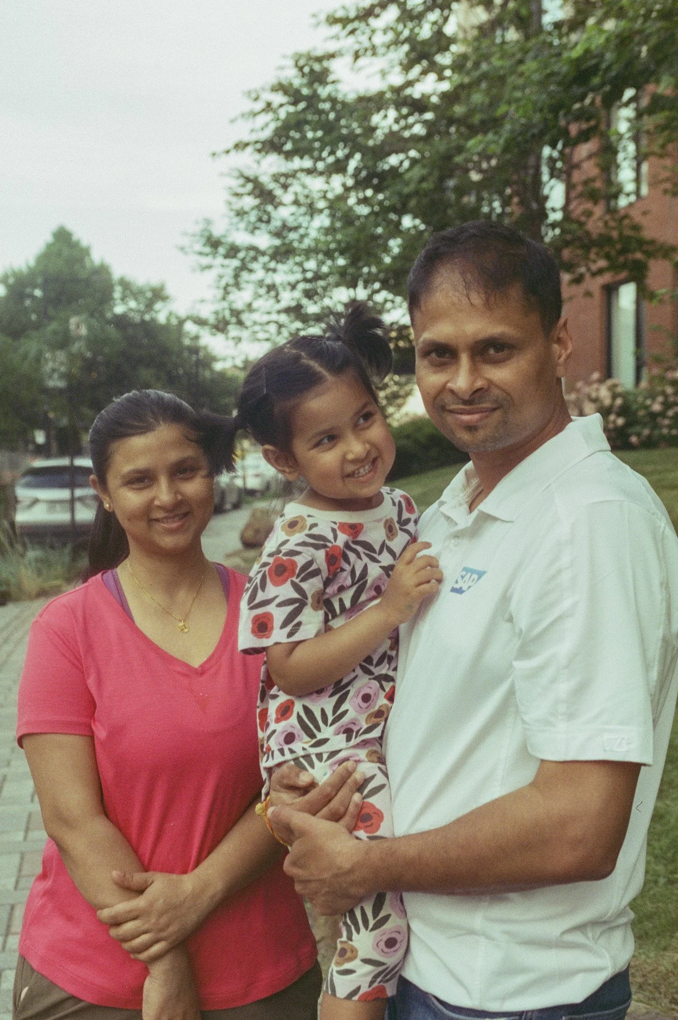A family of three people, a man, a woman, and a young girl, standing outdoors on a sidewalk in front of greenery and a brick building. The man is holding the girl, and all are smiling and looking at the camera.