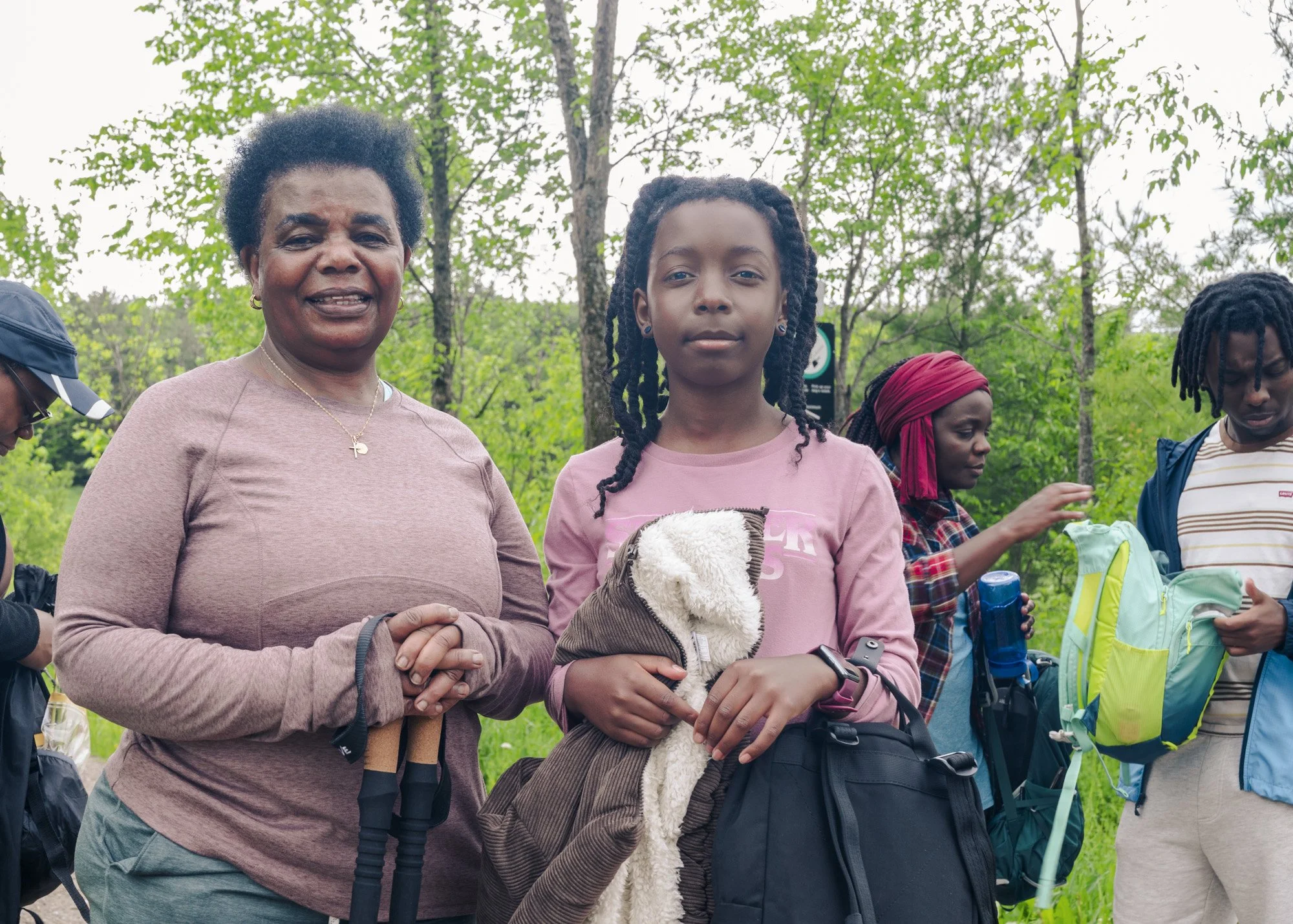 A group of people outdoors in a wooded area during daytime. Central figures include an older woman with short curly hair wearing a pink top and a young girl with braided hair wearing a pink shirt holding a stuffed animal. Other individuals in the bac