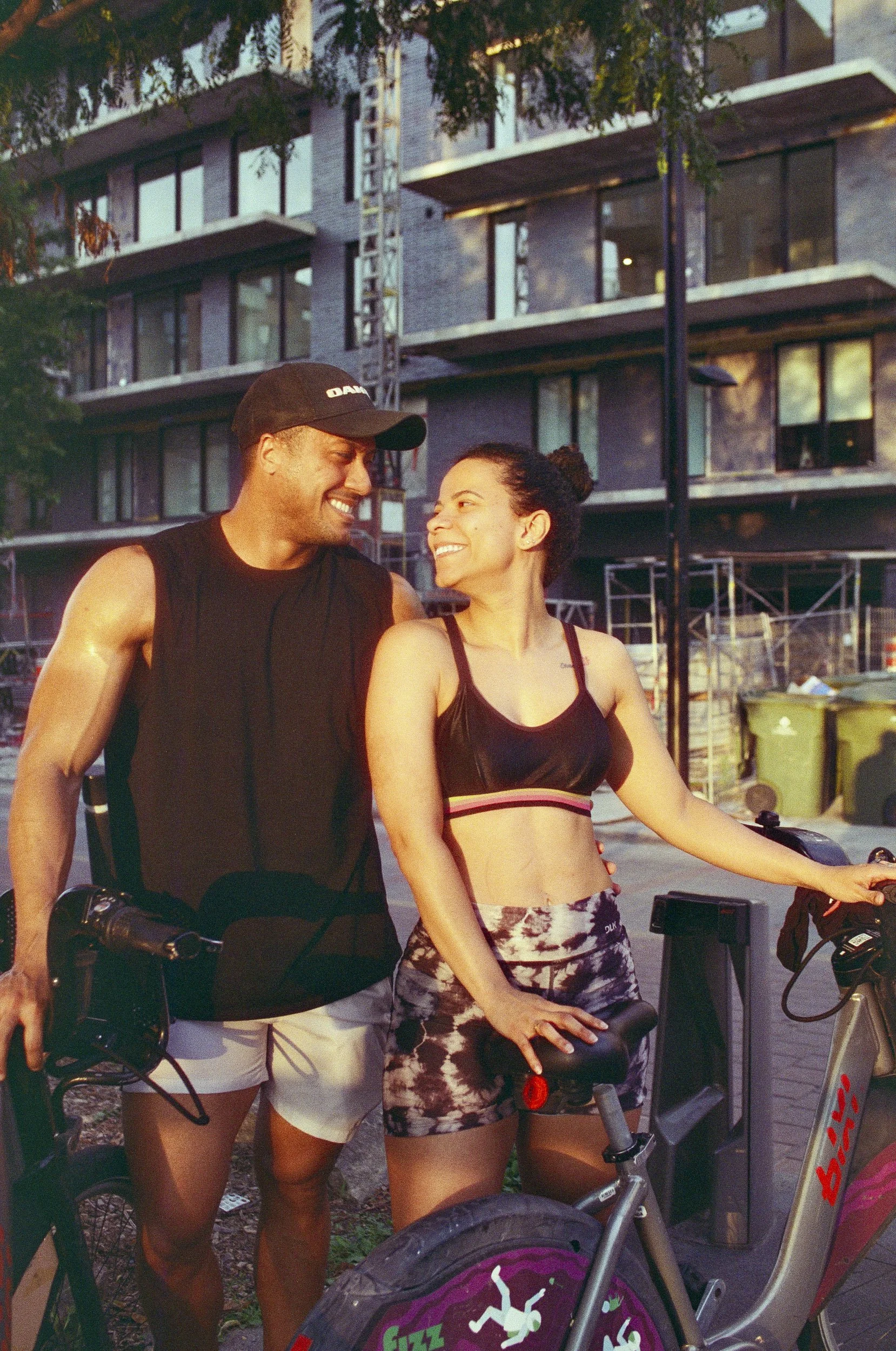 A man and a woman smiling at each other while standing next to bicycles in front of a modern apartment building during daytime. The man is wearing a black sleeveless shirt and a black cap, and the woman is wearing a black sports bra and patterned sho