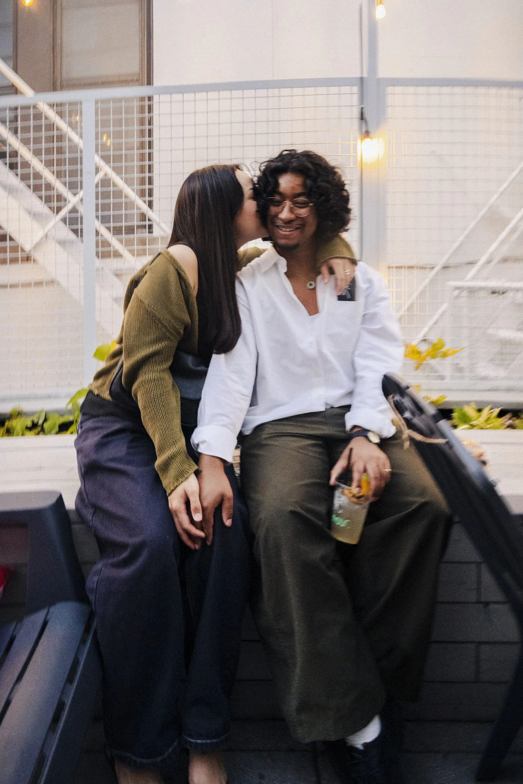 A woman is kissing a man on the cheek while he smiles, seated on a bench in a modern indoor space. She has long dark hair and is wearing a green jacket and dark pants. He has curly hair, glasses, and is wearing a white shirt and dark pants, holding a
