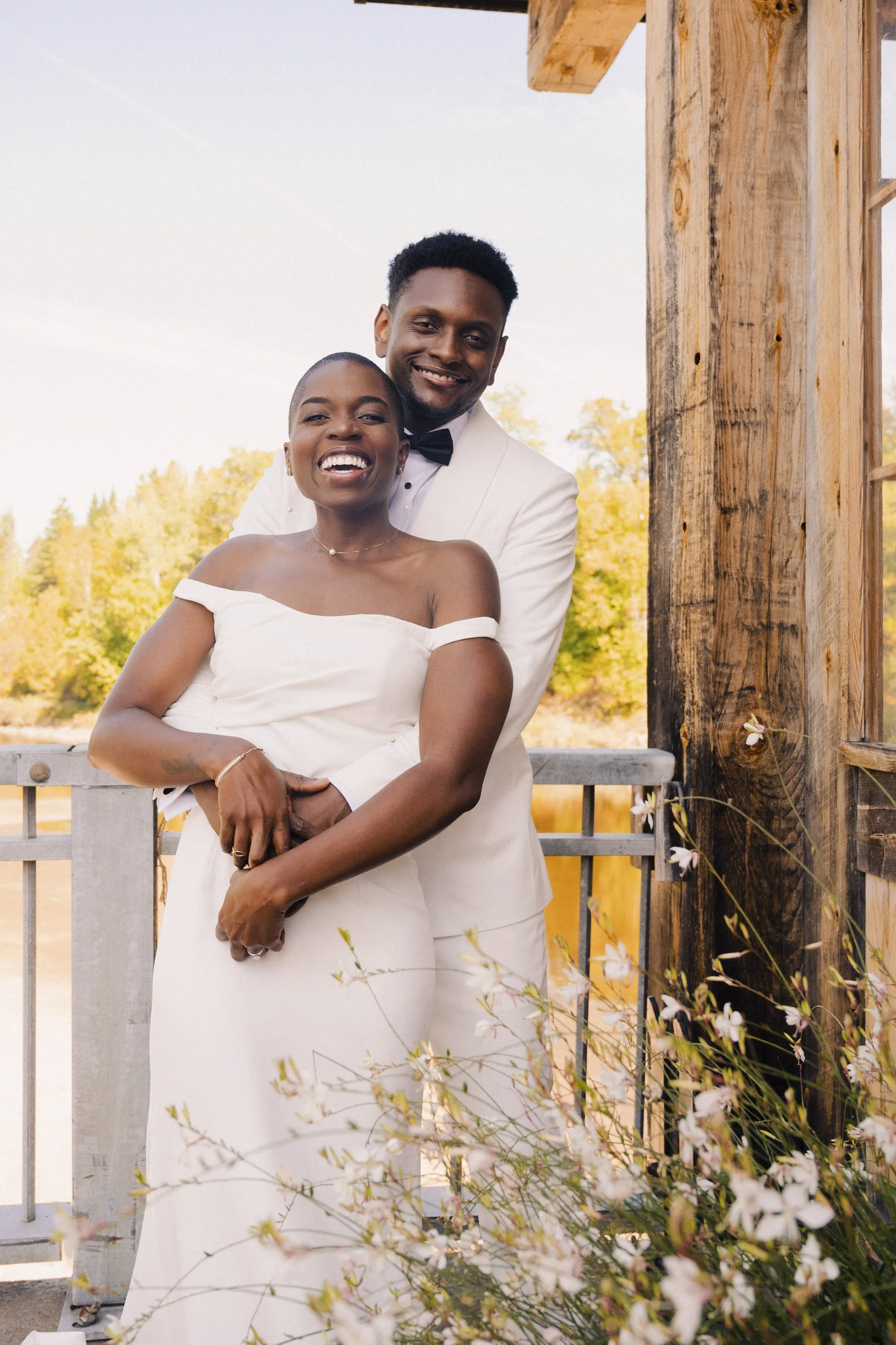 A smiling woman in a white off-shoulder dress and a man in a white tuxedo with a black bow tie, standing outdoors during sunset near a wooden structure and blooming flowers.