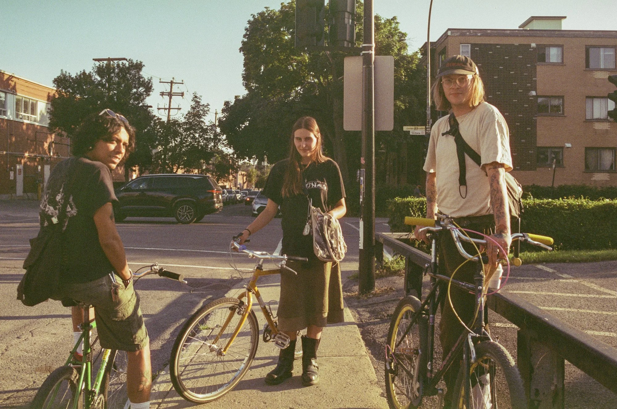 Three young women with bicycles standing on a sidewalk in a city neighborhood during late afternoon or early evening with the sun setting. Two women are near the street, one holding a yellow bicycle, and the other standing behind a parked black bicyc