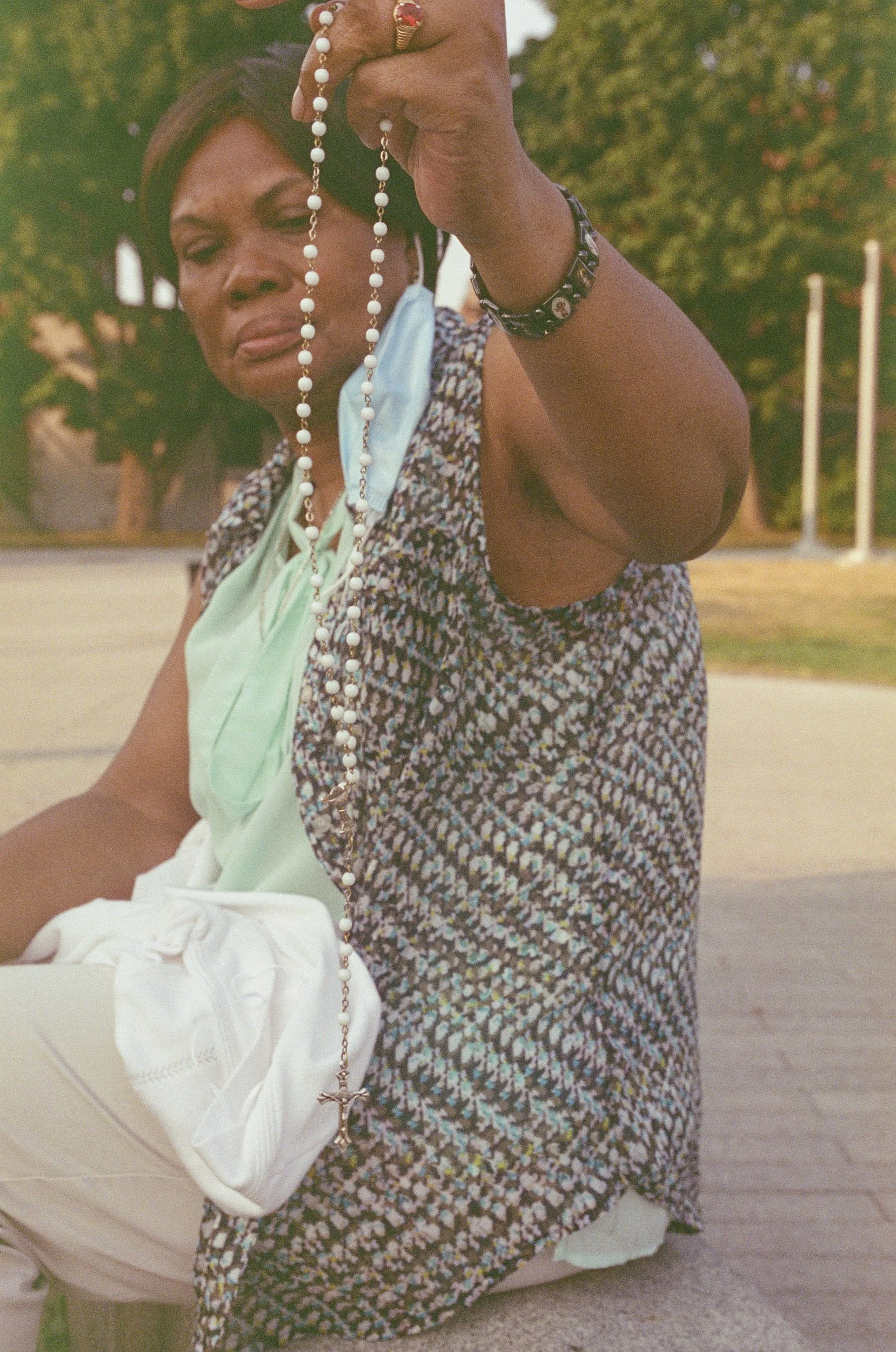 A woman outdoors holding a rosary with white beads and a crucifix, wearing a wristwatch and a patterned dress, with trees and a park in the background.