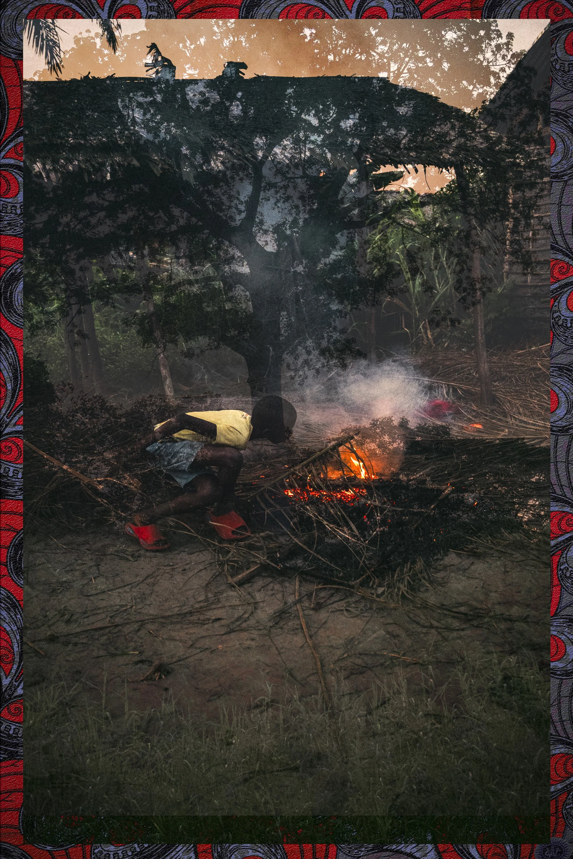 A person in a yellow shirt and red shoes is crouching near a small fire on the ground in a wooded area during dusk or evening. There are trees and smoke visible in the background.