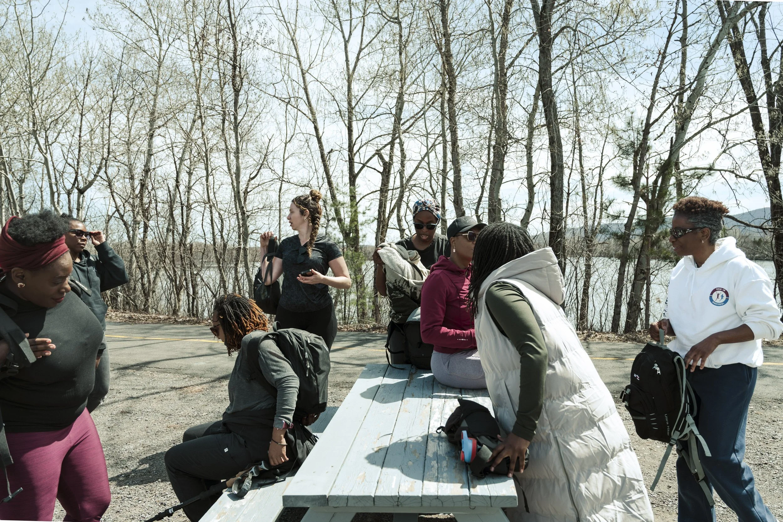 Group of diverse women gathered outdoors around a white picnic table, some standing and some seated, with leafless trees in the background.