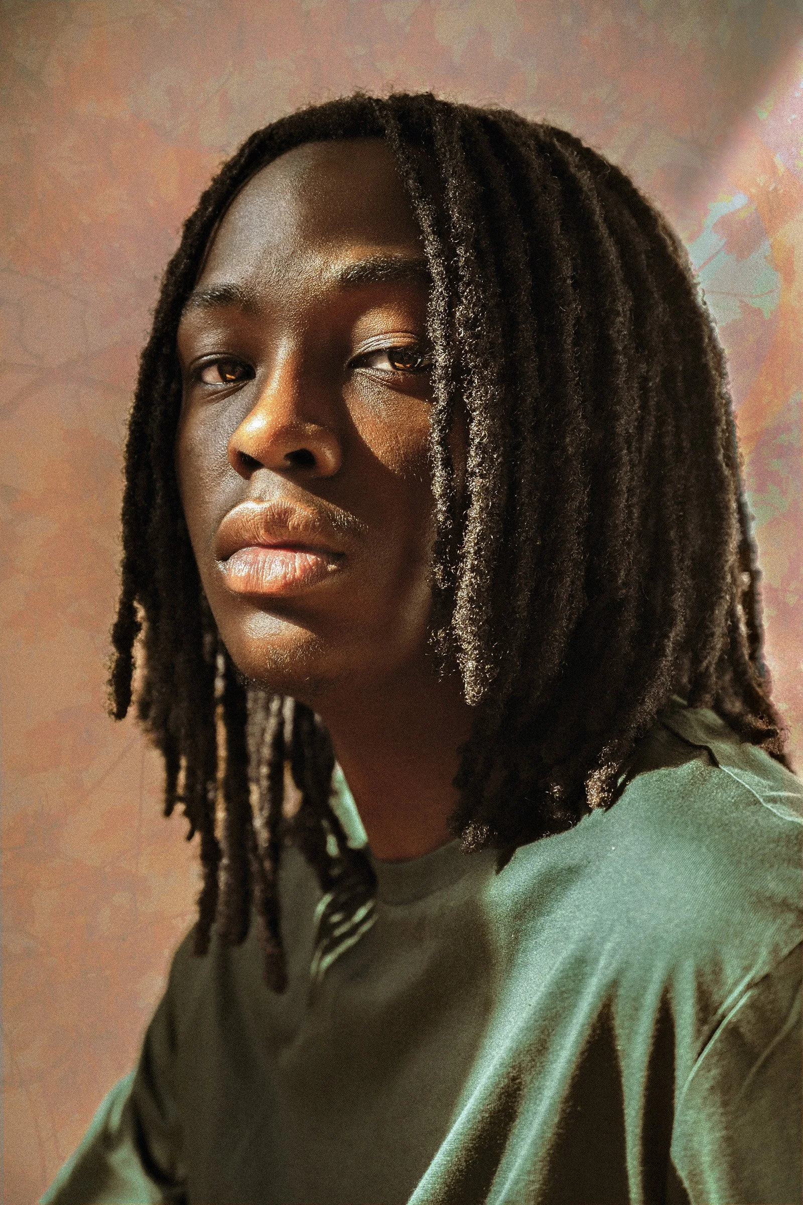 Close-up of a woman with dark skin and dreadlocks, wearing a green sweatshirt, looking at the camera with a neutral expression.