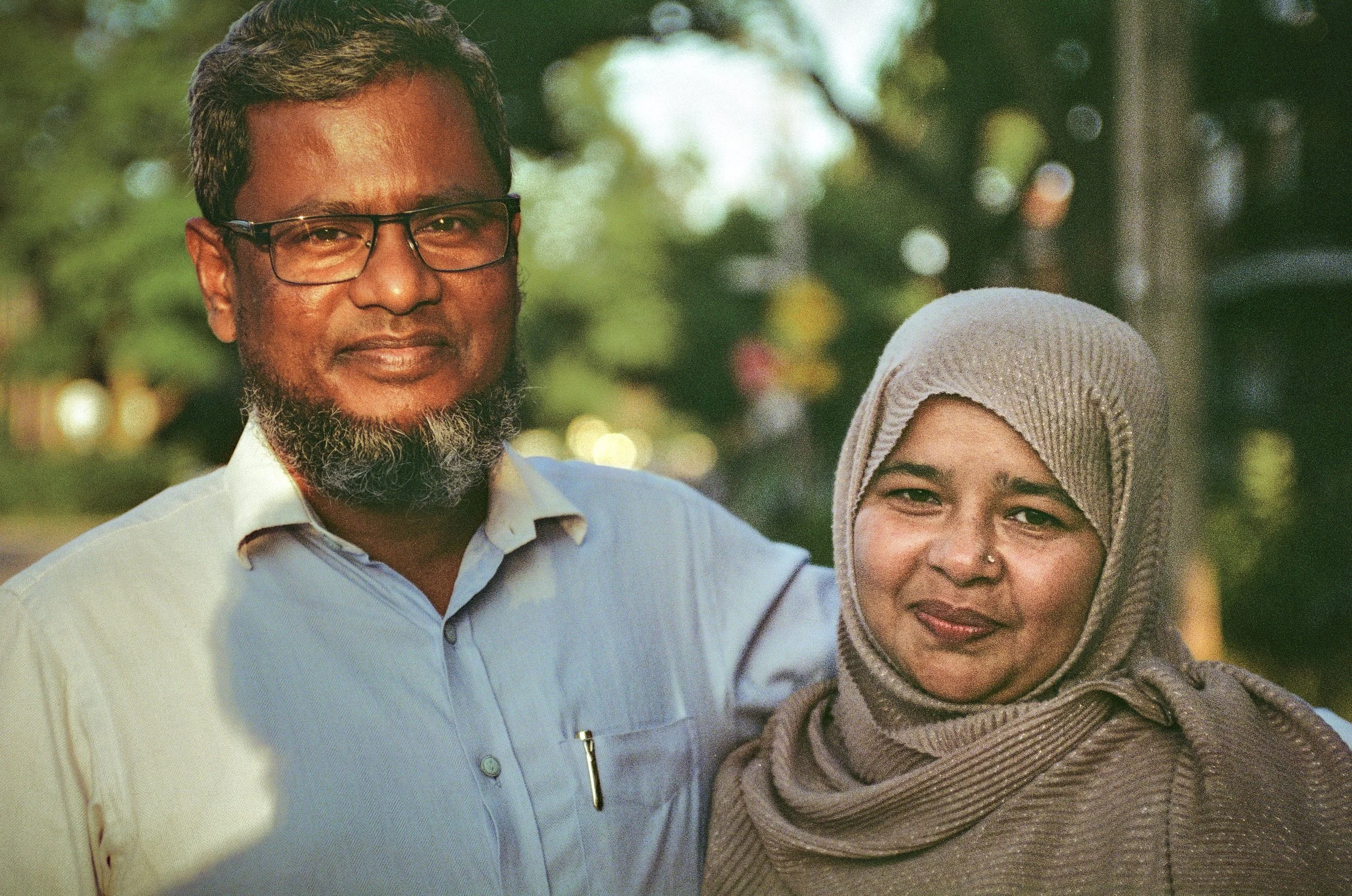 A man with glasses and a beard smiling, standing outdoors with a woman wearing a hijab, both looking at the camera.