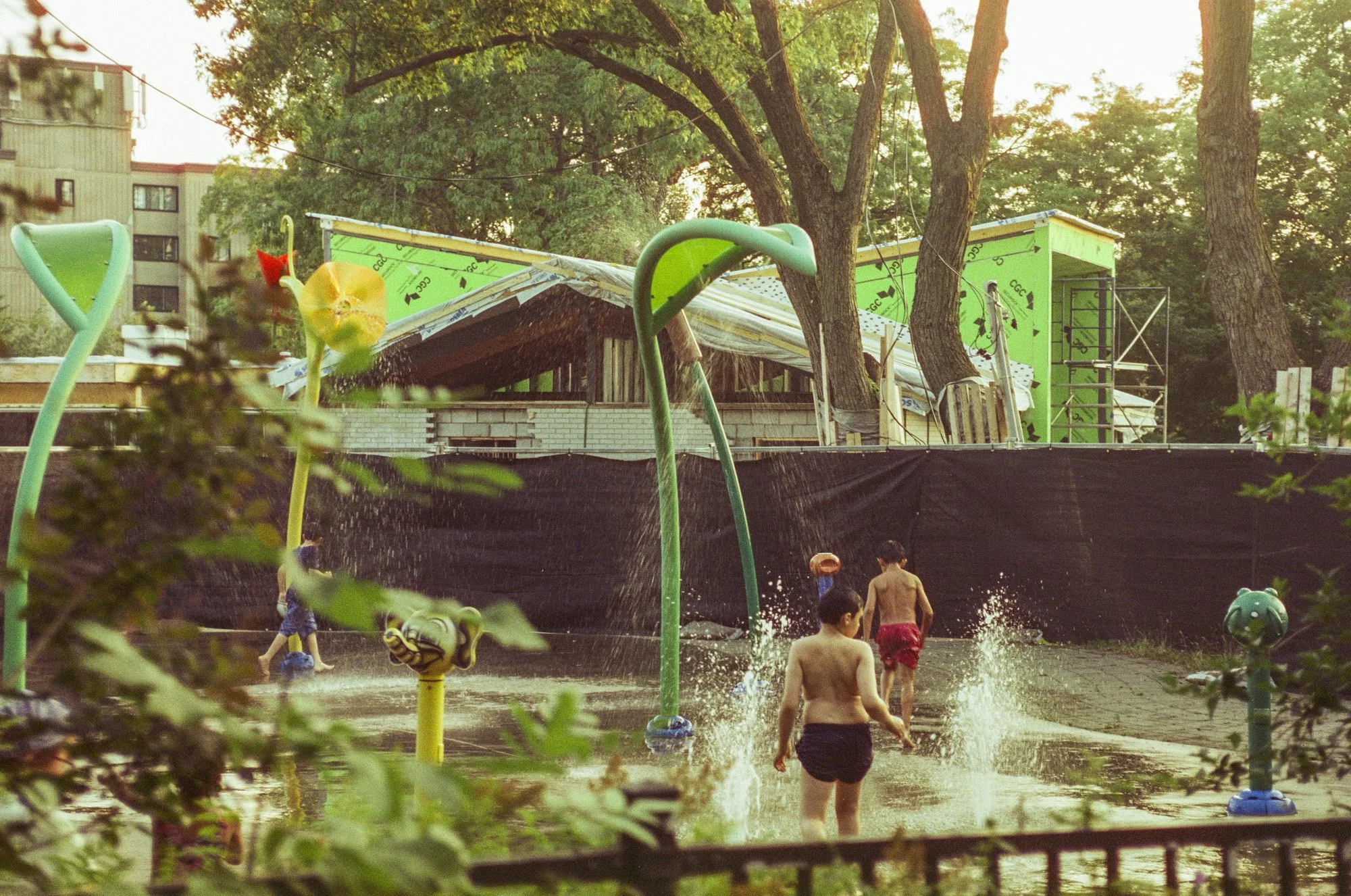 Children playing at a splash pad with water sprinklers and geysers, trees in the background, and some construction or renovation happening on a building.