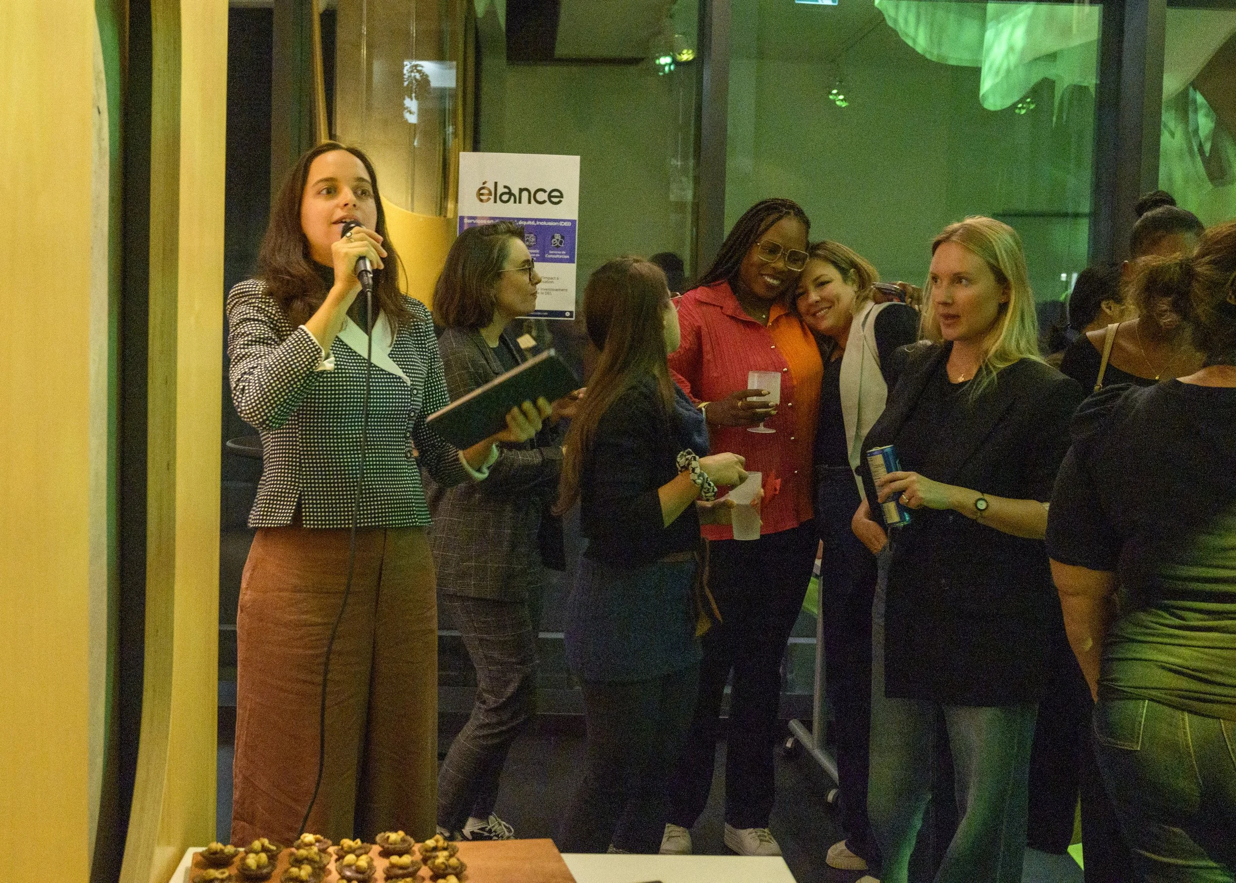 Group of women at a social or networking event, some holding drinks, standing near a green wall with a large green decorative element, and a table with snacks or desserts in the foreground.
