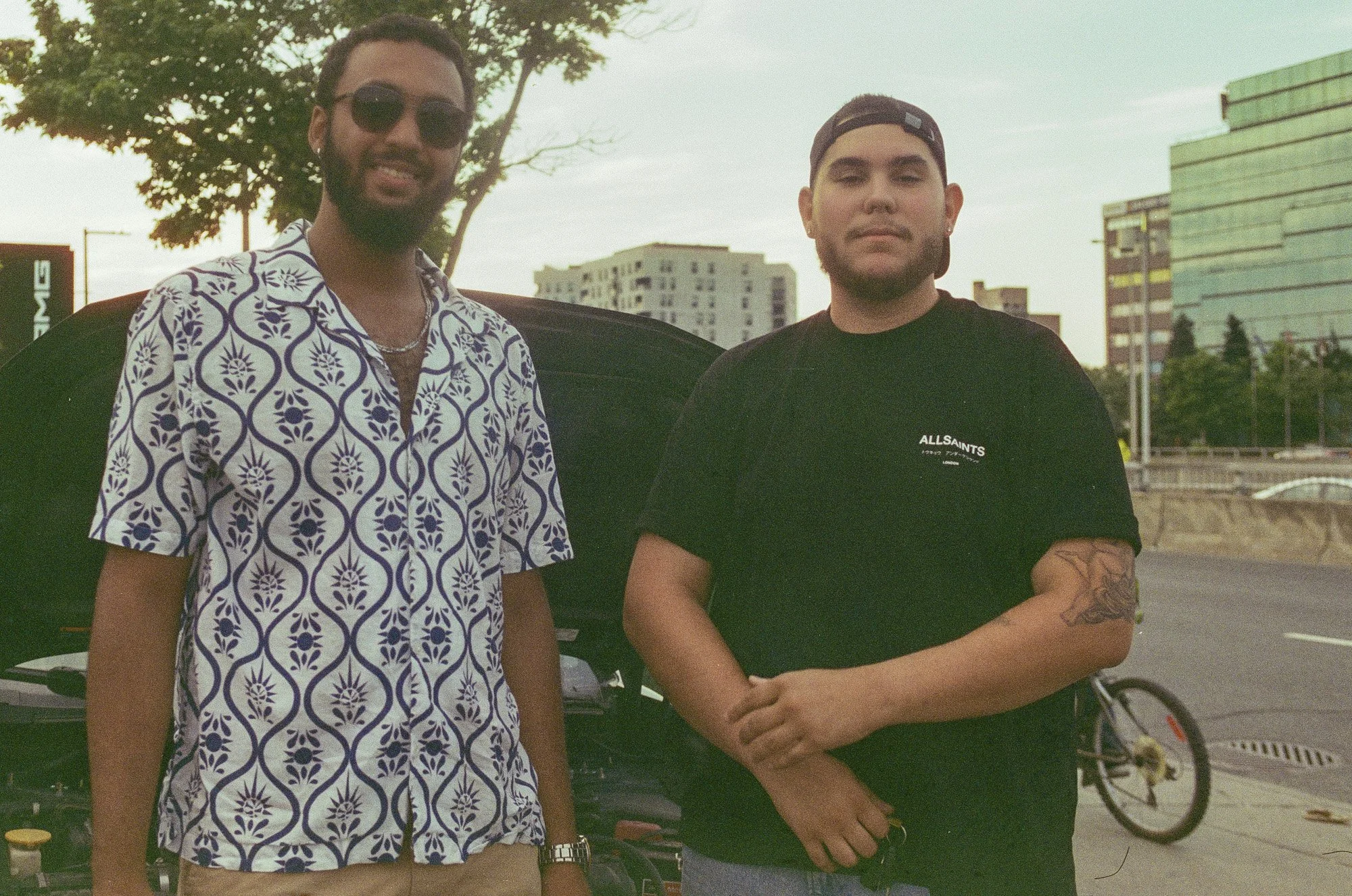Two young men standing outdoors in urban setting during daytime, with buildings and trees in the background. One wearing sunglasses and a patterned shirt, the other wearing a black T-shirt and a backward cap.