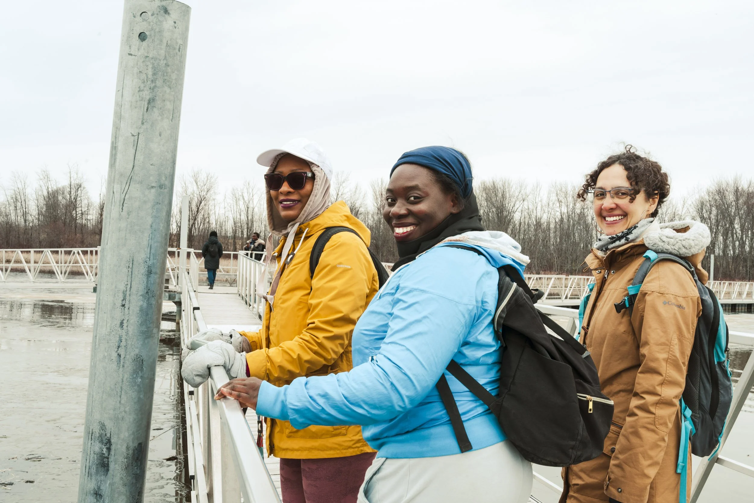 Three women standing on a dock, wearing jackets and backpacks, looking towards the camera in a chilly outdoor setting during overcast weather.