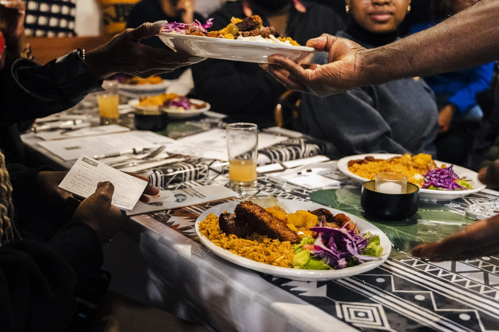 People at a restaurant table sharing plates of colorful grilled food, rice, and vegetables, with glasses of orange juice on a patterned tablecloth.