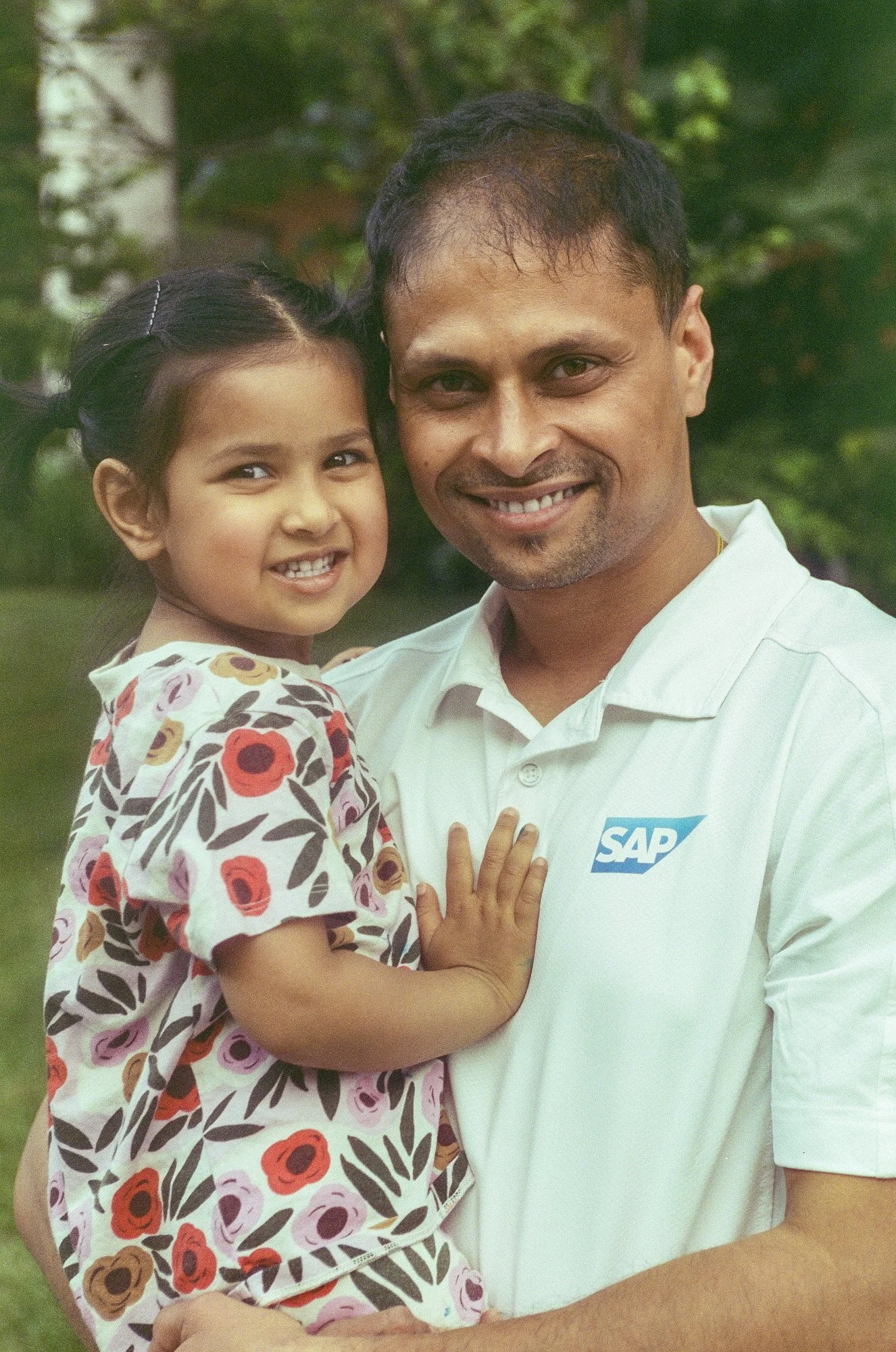 A man and a young girl are smiling and hugging outdoors, with greenery in the background. The man is wearing a white polo shirt with an SAP logo.