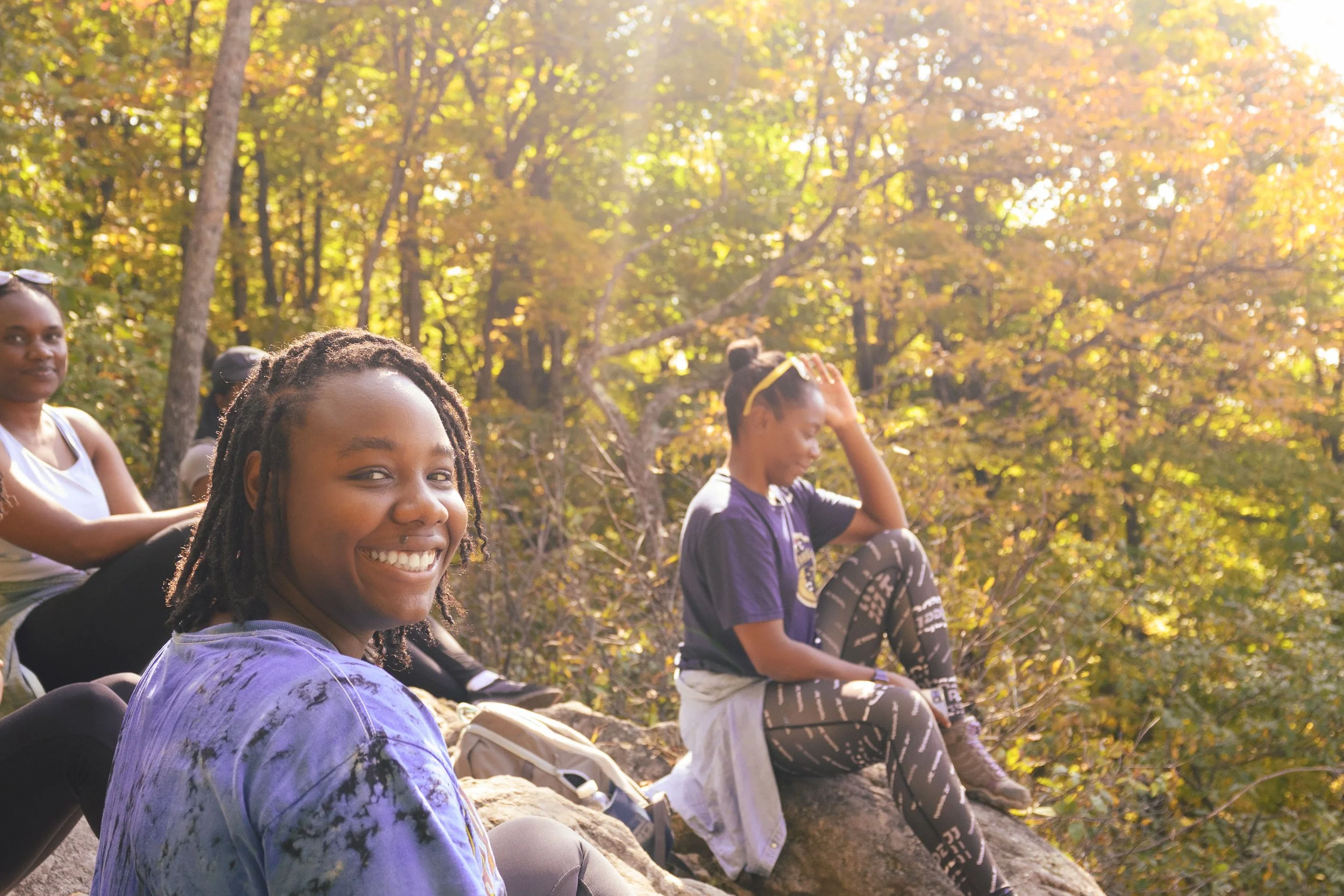 A group of diverse young women sitting outdoors on a fall day surrounded by yellow and orange leaves on trees, enjoying nature.