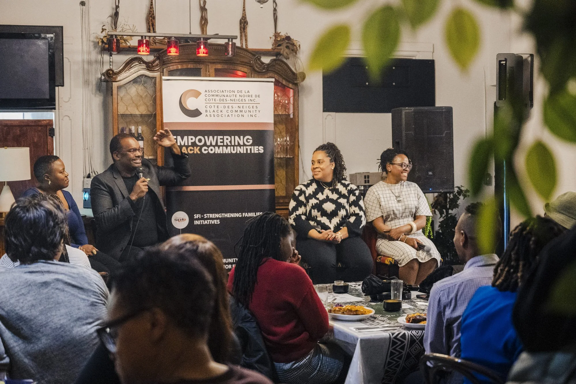 A group of people gathered in a room listening to a speaker. The speaker, a man in a suit, is holding a microphone and gesturing with his hand. Behind him is a banner that reads, 'Empowering Black Communities,' and the banner belongs to the Cote-des-