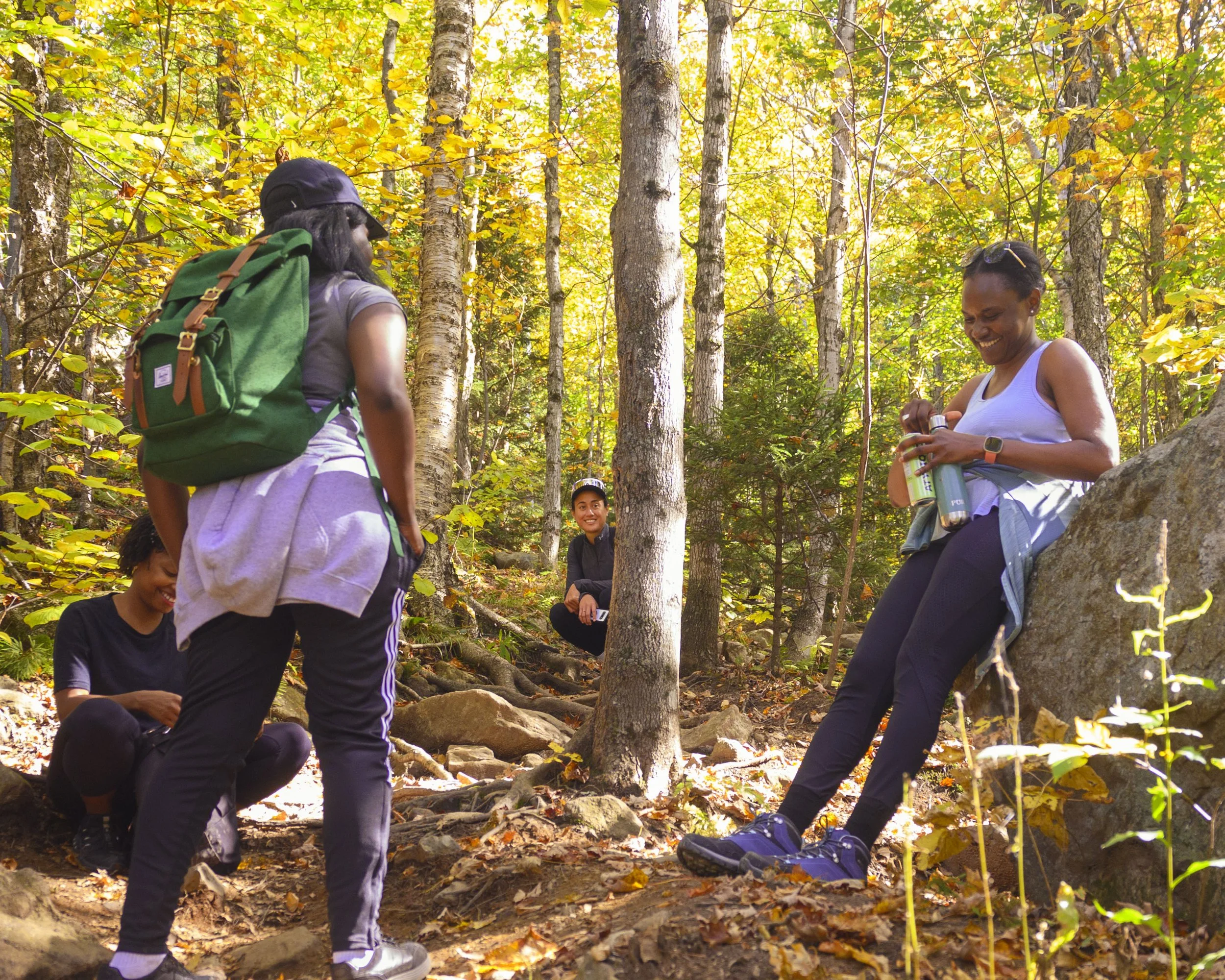 Group of hikers in a forest with trees shedding yellow leaves, some standing and some sitting on rocks and logs, preparing for a hike.