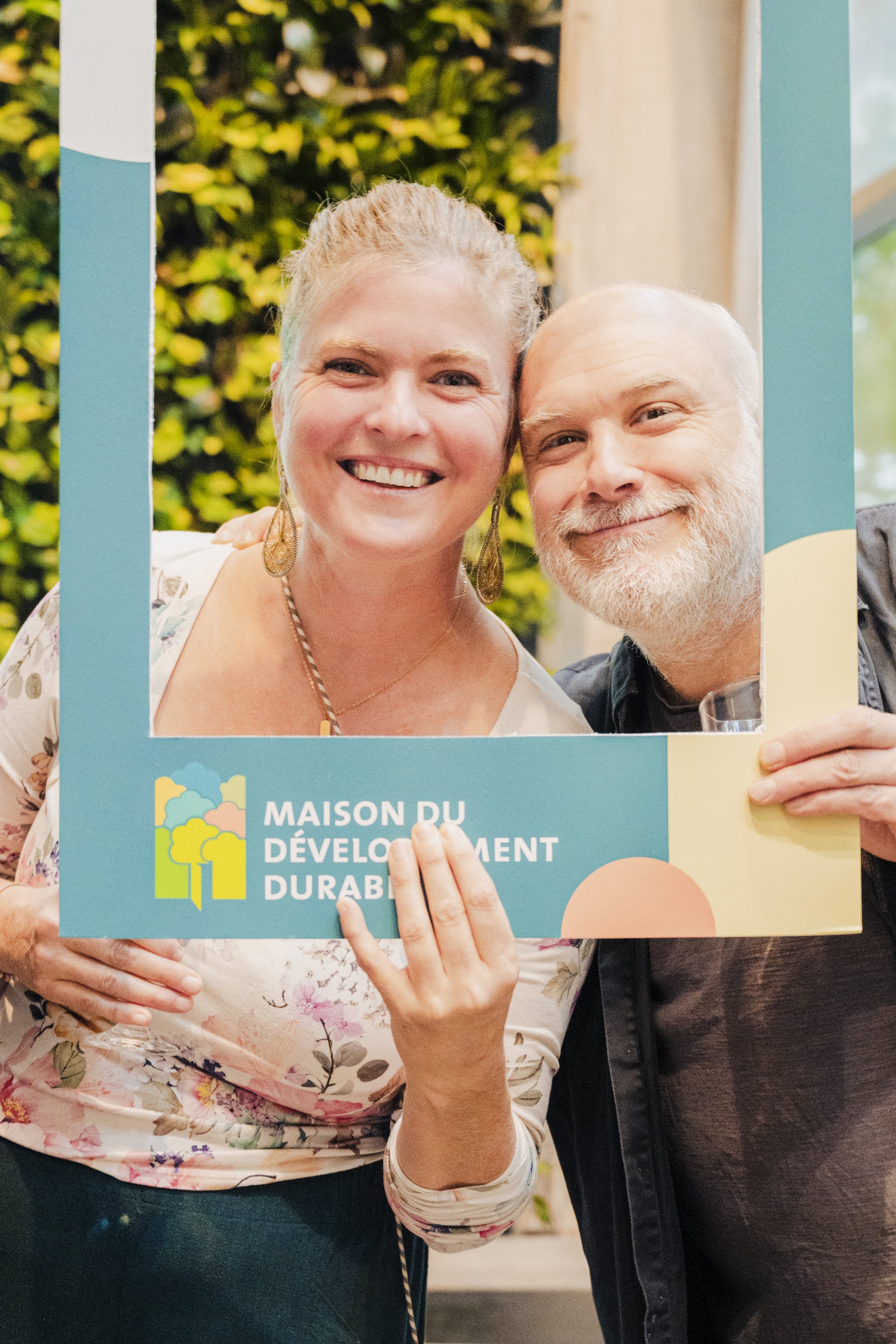 A woman and a man smiling and holding a sign that reads 'MAISON DU DÉVELOPPEMENT DURABLE'. The woman is wearing earrings, a floral top, and has her hair pulled back. The man has a beard, is smiling, and is holding a glass. They are standing indoors w