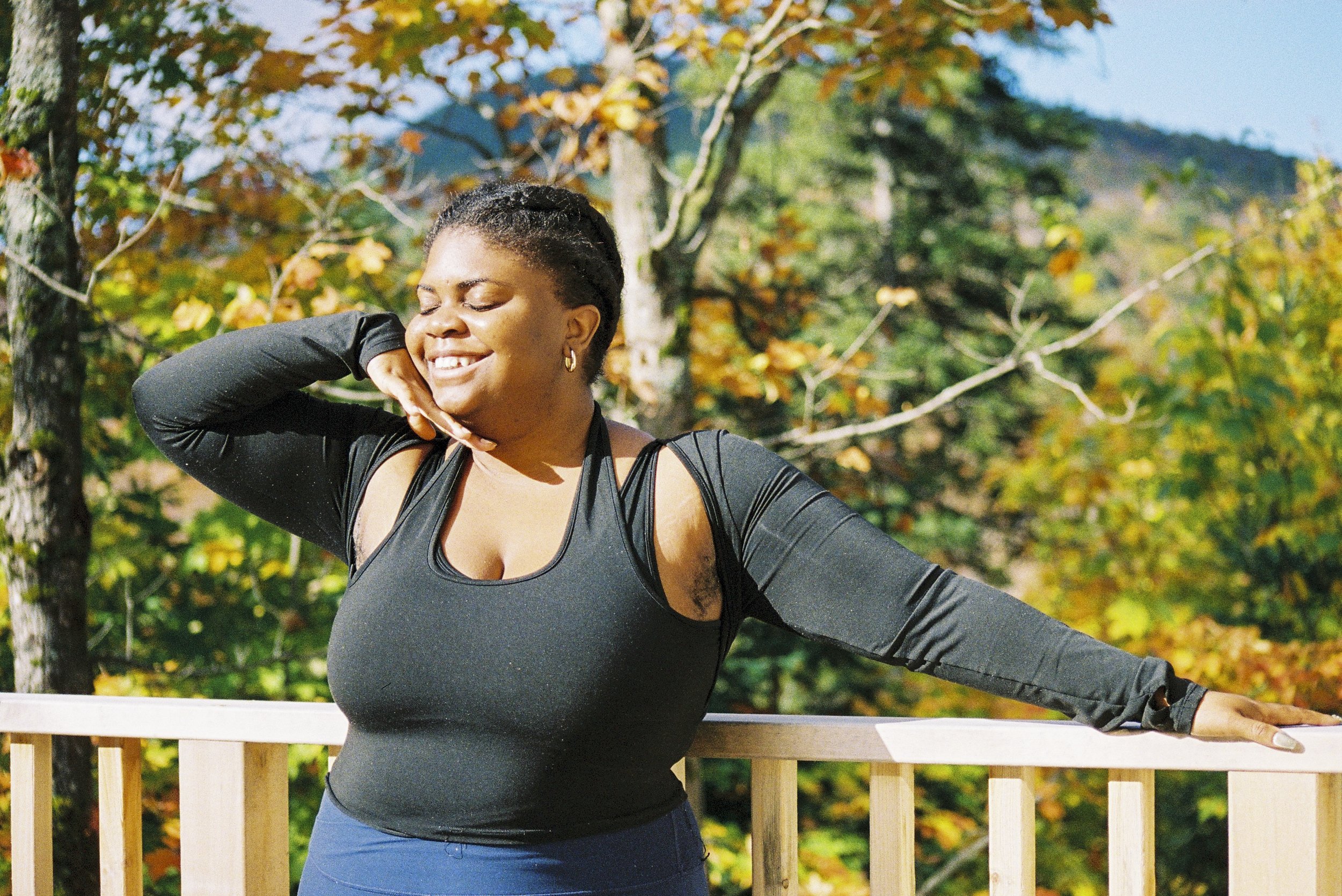 A woman in athletic clothing smiling with closed eyes outdoors, surrounded by trees with autumn leaves, leaning on a wooden railing.