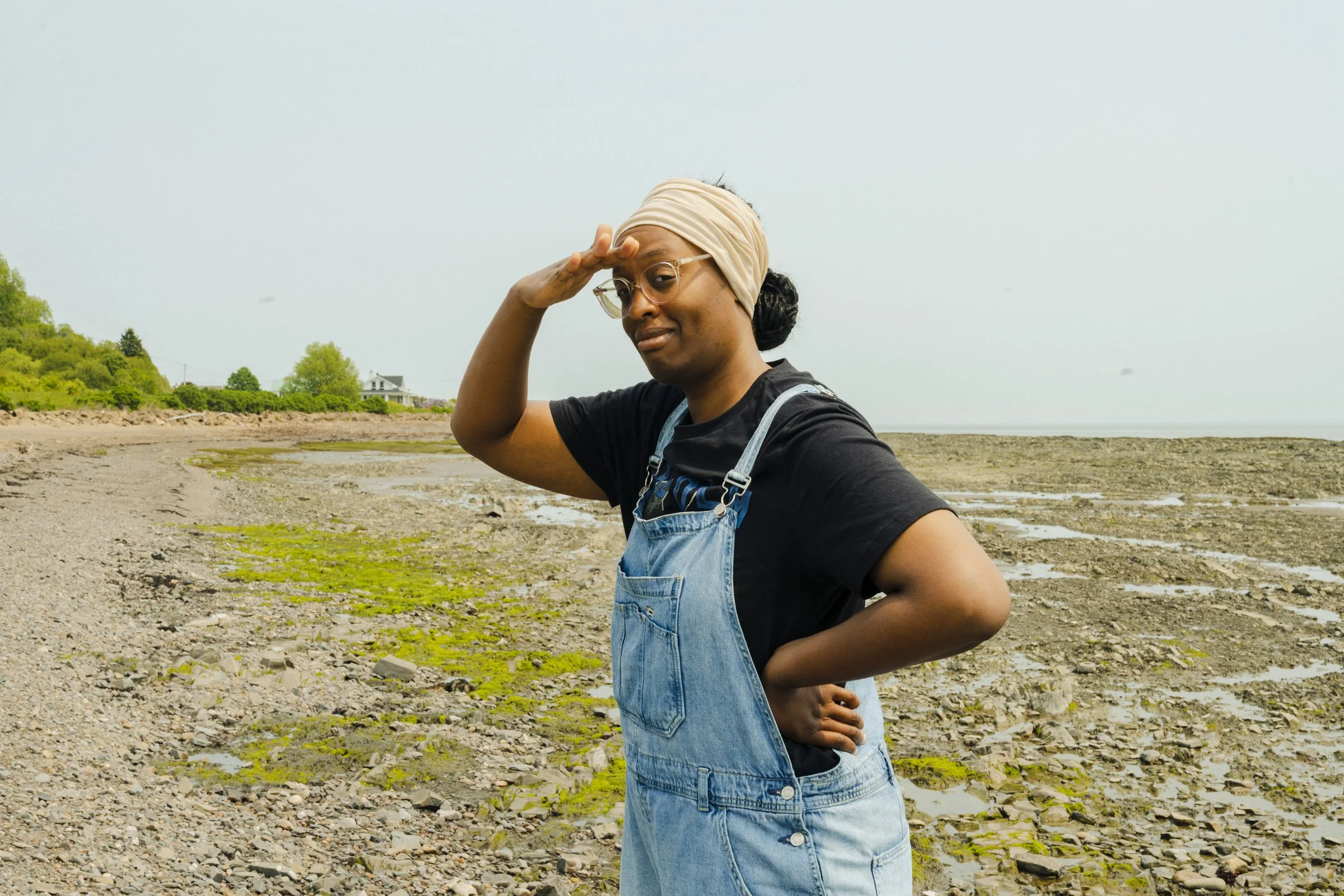 Woman standing on a rocky beach with her hand raised to her forehead, wearing glasses, a beige head wrap, a black t-shirt, and denim overalls, looking towards the camera with a slight smile.