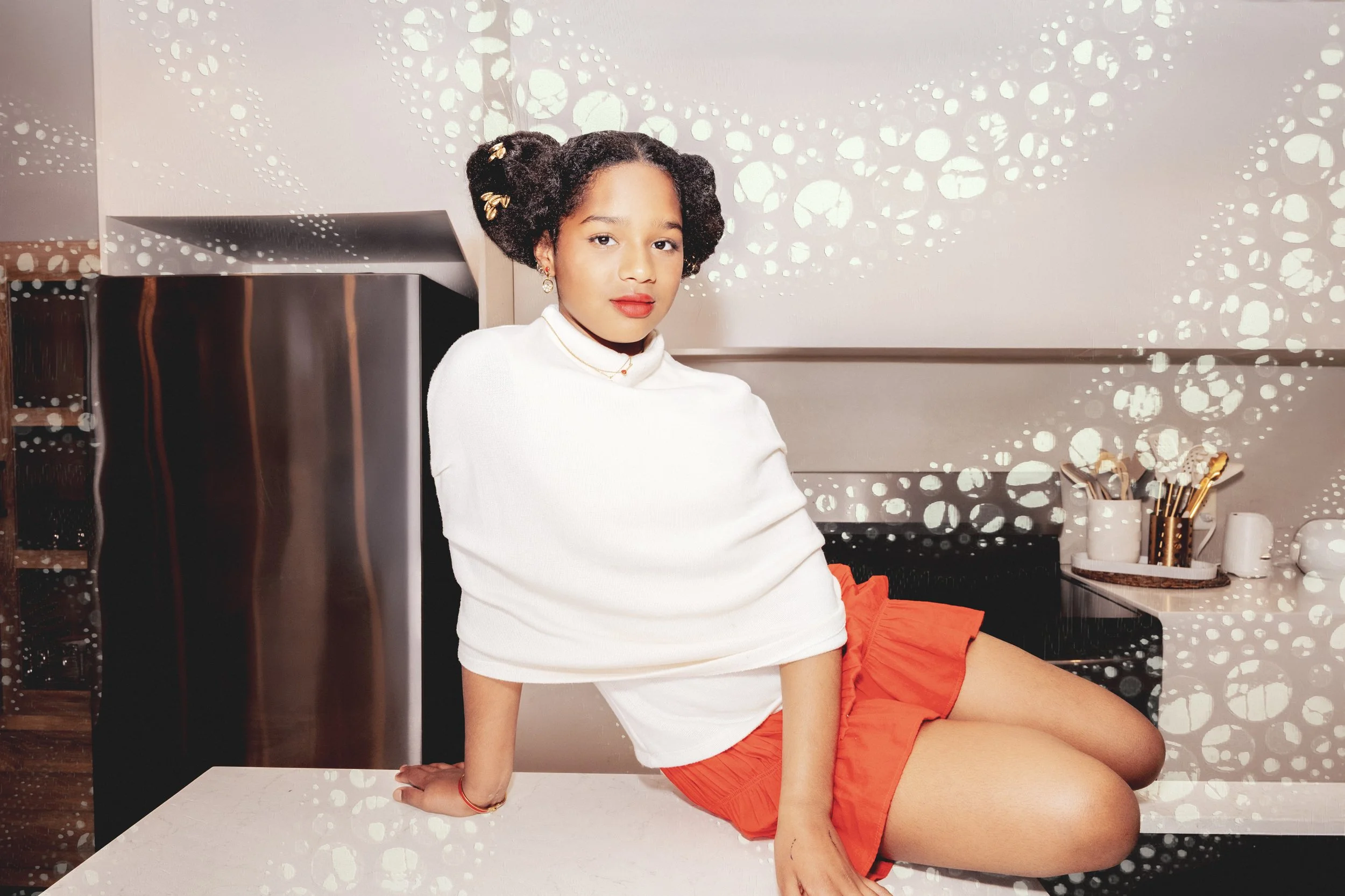 A young girl sitting on a kitchen countertop, wearing a white turtleneck and orange shorts, with short curly hair decorated with hair clips, and looking at the camera.