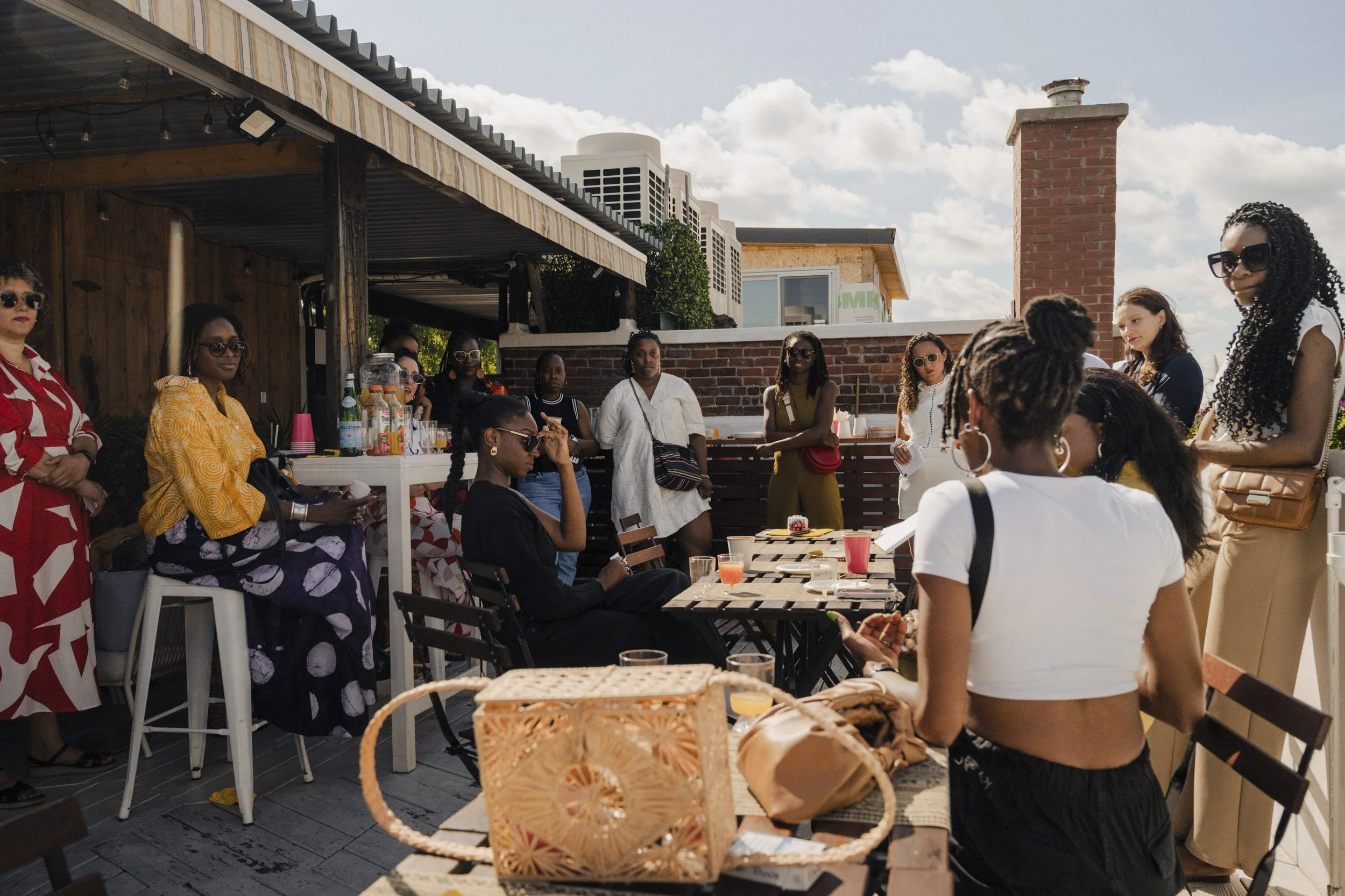 A group of people gathered outdoors on a rooftop terrace, enjoying a social event with drinks and snacks on a sunny day. Some are seated, while others are standing and conversing.