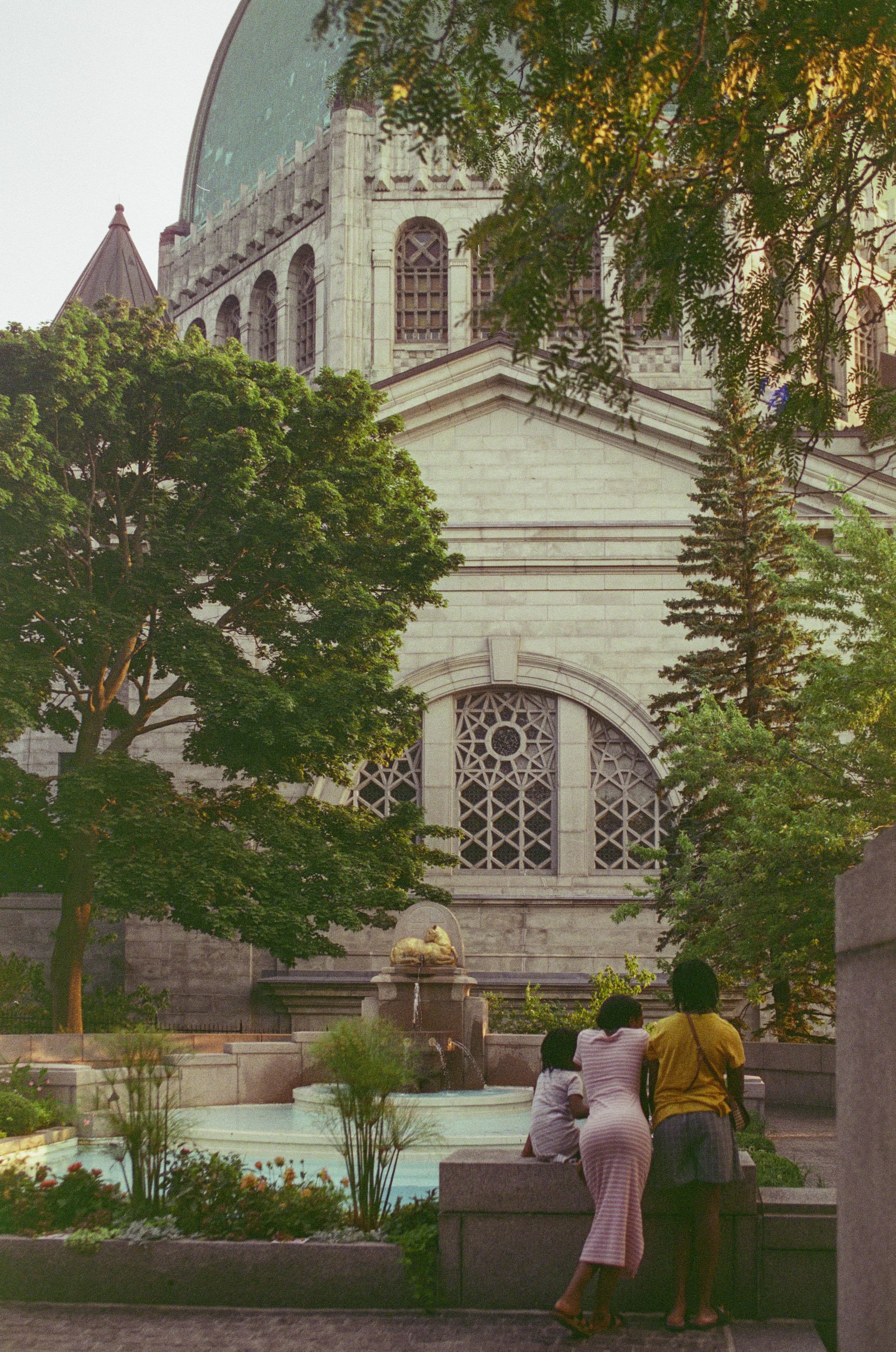 A park scene with a large stone building in the background, which appears to be a church or cathedral, with trees framing the scene. In the foreground, three people are sitting on a bench near a fountain, with two of them engaged in conversation and 