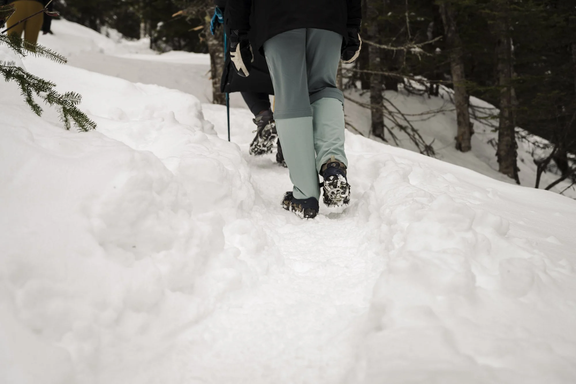 Person walking in snowy forest, wearing winter gear and snow boots, with snow-covered trail and trees in background.