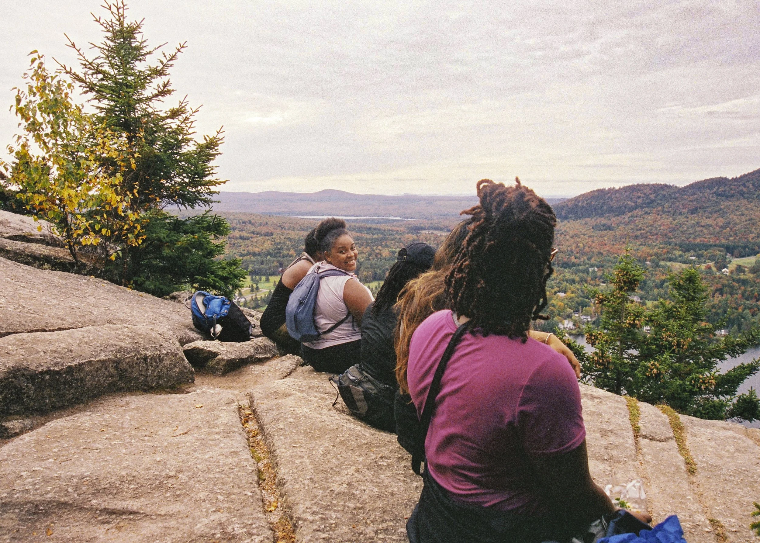 A group of people sitting on rocks, overlooking a scenic landscape with mountains, trees, and a cloudy sky, during what appears to be autumn.