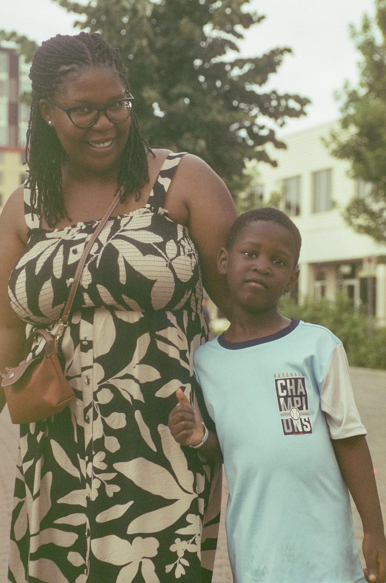 A woman with glasses and earrings, wearing a black and white floral dress, and a boy in a light blue t-shirt with sports graphics, standing outdoors in front of trees and buildings, smiling and making a thumbs-up gesture.