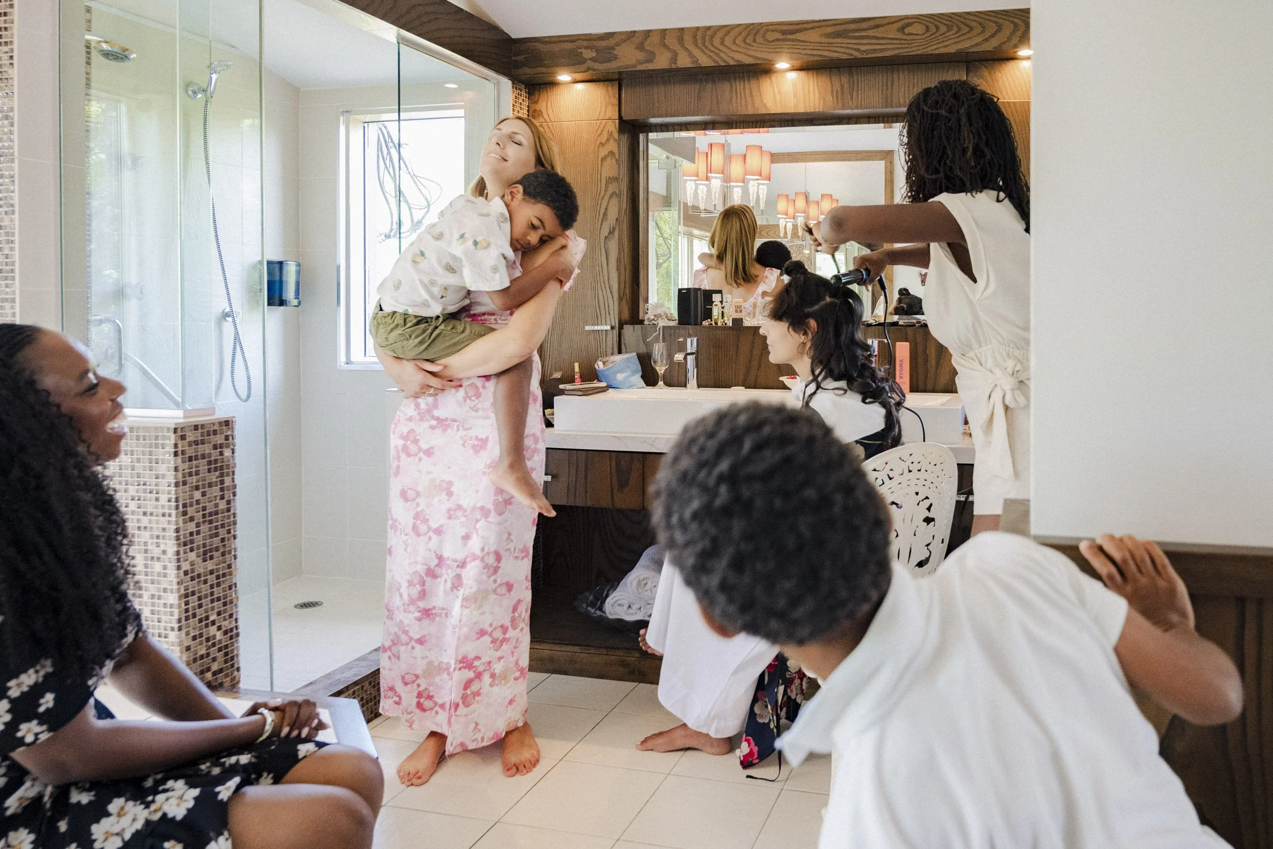 A woman is holding a child on her shoulder while surrounded by others in a room, with a woman styling another woman's hair. The scene appears to be at a party or gathering, with a bathroom and mirror visible in the background.
