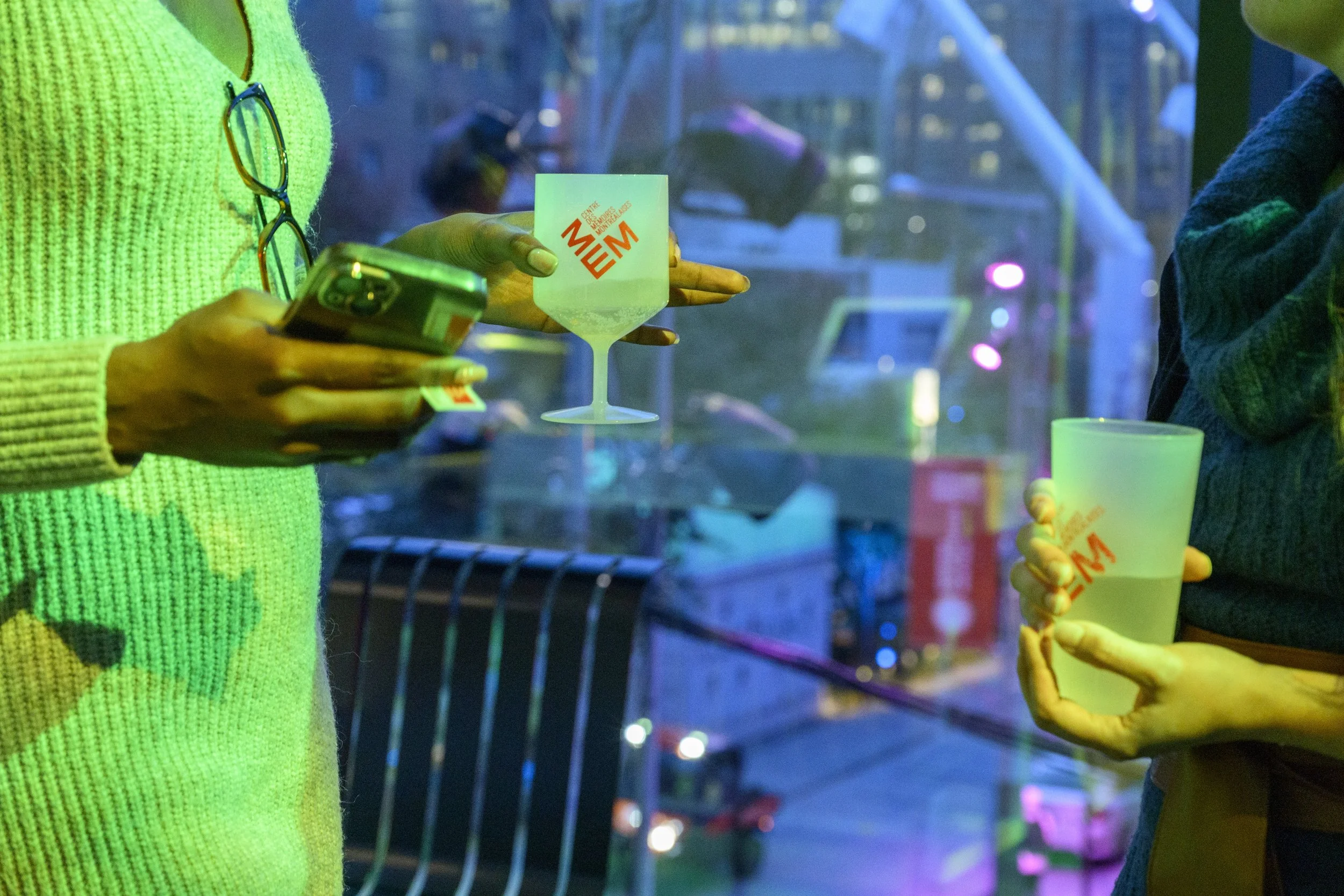 Two people holding glasses with the URL 'MEM' printed on them. One person is wearing a neon green sweater with glasses hanging on the collar, and the other person is wearing a dark sweater, holding a similar glass. The background features colorful, n