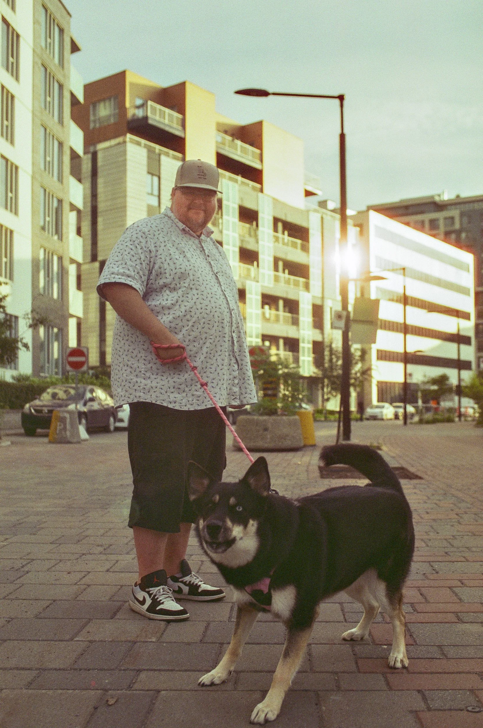 A man standing on a city sidewalk with a Siberian Husky dog, modern buildings in the background, and the sun setting.