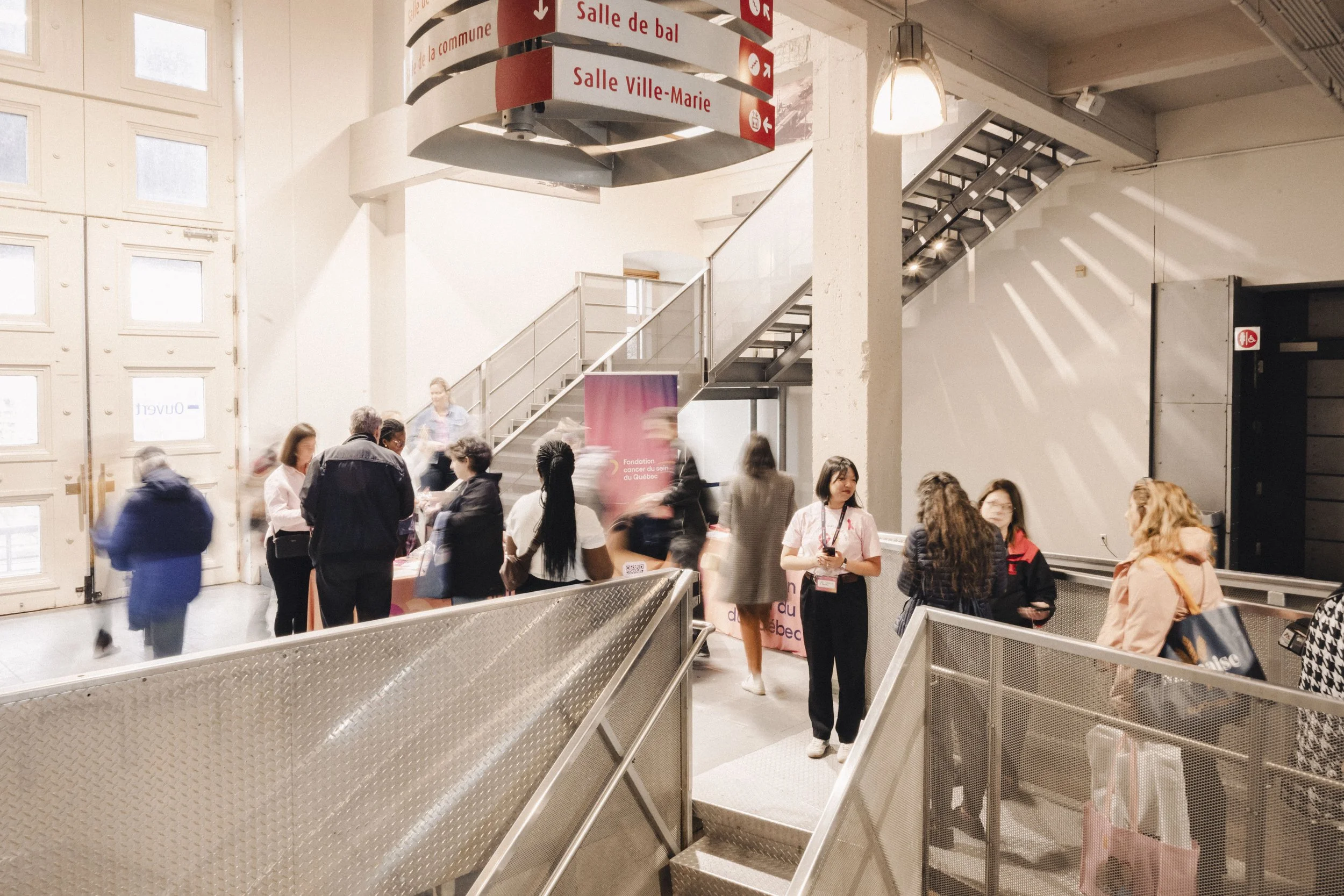 A busy indoor event with people gathered around a table near a stairway. There are signs in French hanging from the ceiling indicating directions to different rooms. A banner for a cancer foundation is visible, and some people are engaged in conversa