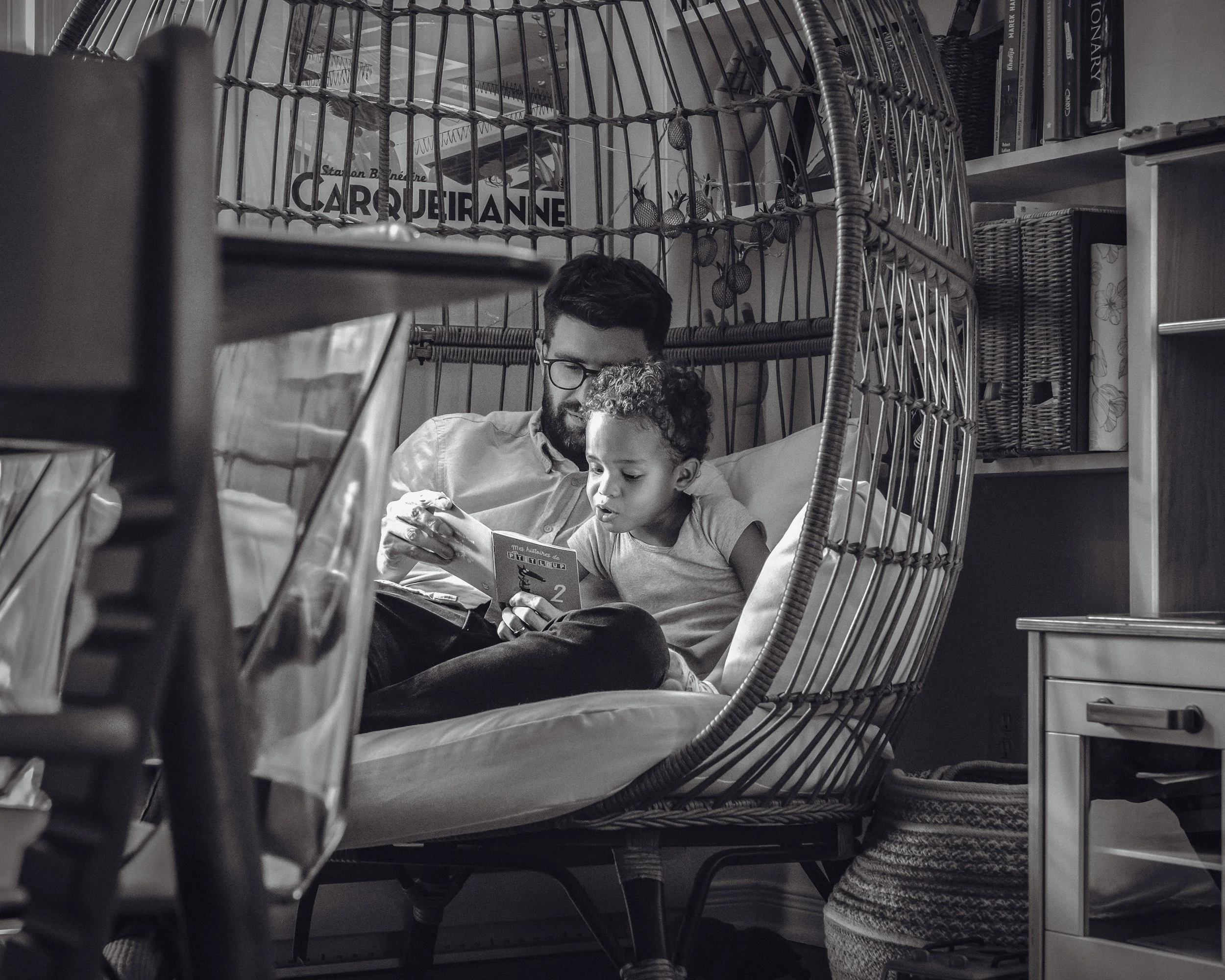 A man with glasses and a beard and a young boy sit together on a bed, looking at a device. The background shows a bookshelf with books and decorative items, a wicker basket, and a library sign that says 'CARROUITAN'. The photo is in black and white.