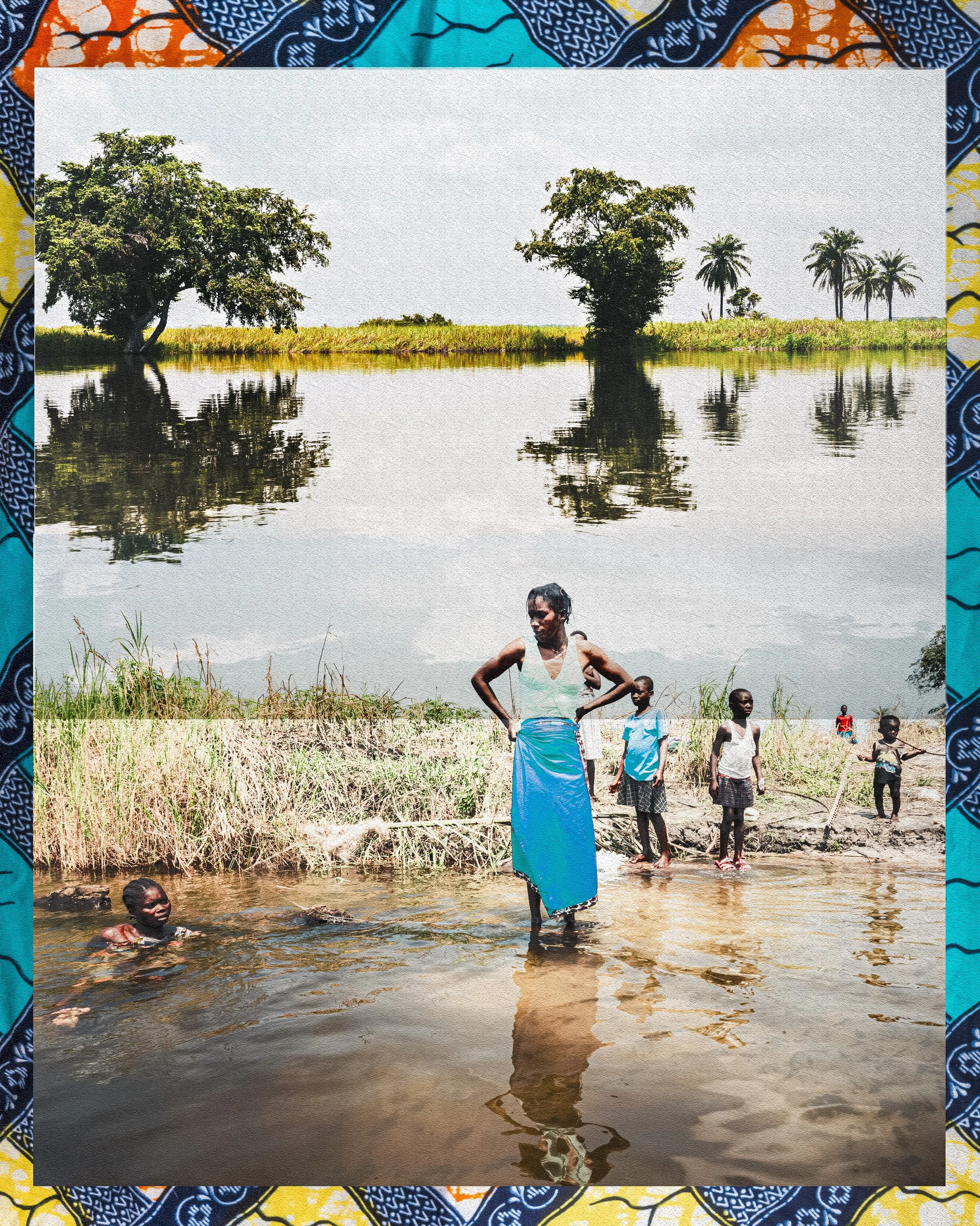A group of people, including women and children, are gathered near a body of water in a rural area. Some are standing in the water while others are on the grassy bank. The background features trees and a landscape with palm trees.