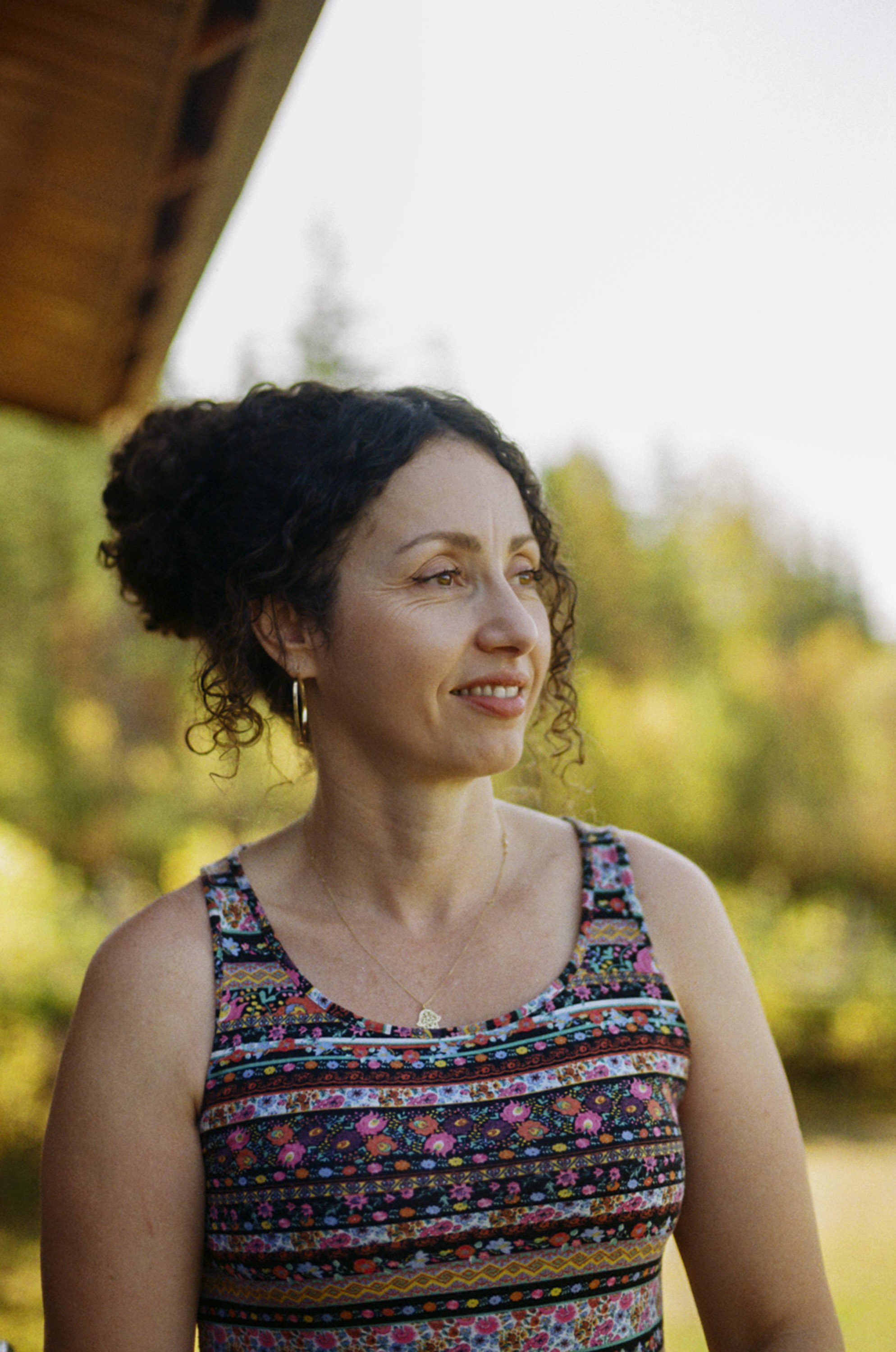 A young woman with curly dark hair standing outdoors, smiling softly, with trees and a bright sky in the background. She is wearing a colorful, patterned sleeveless dress and gold hoop earrings.