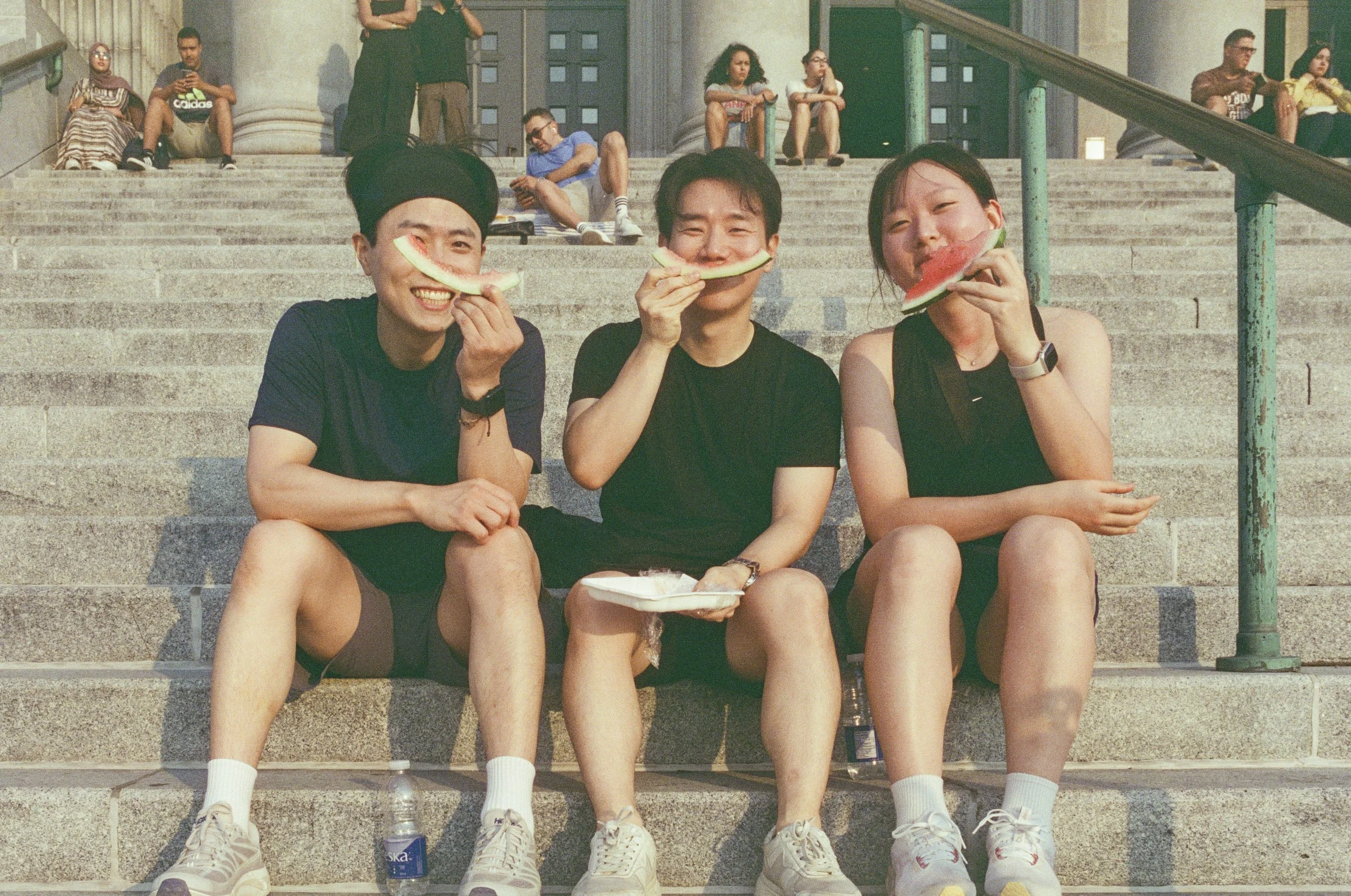 Three young people sitting on steps outside, eating watermelon slices. The person on the left wears a black T-shirt, baseball cap, and sneakers; the middle person wears a black T-shirt and shorts; the person on the right wears a black tank top, sport