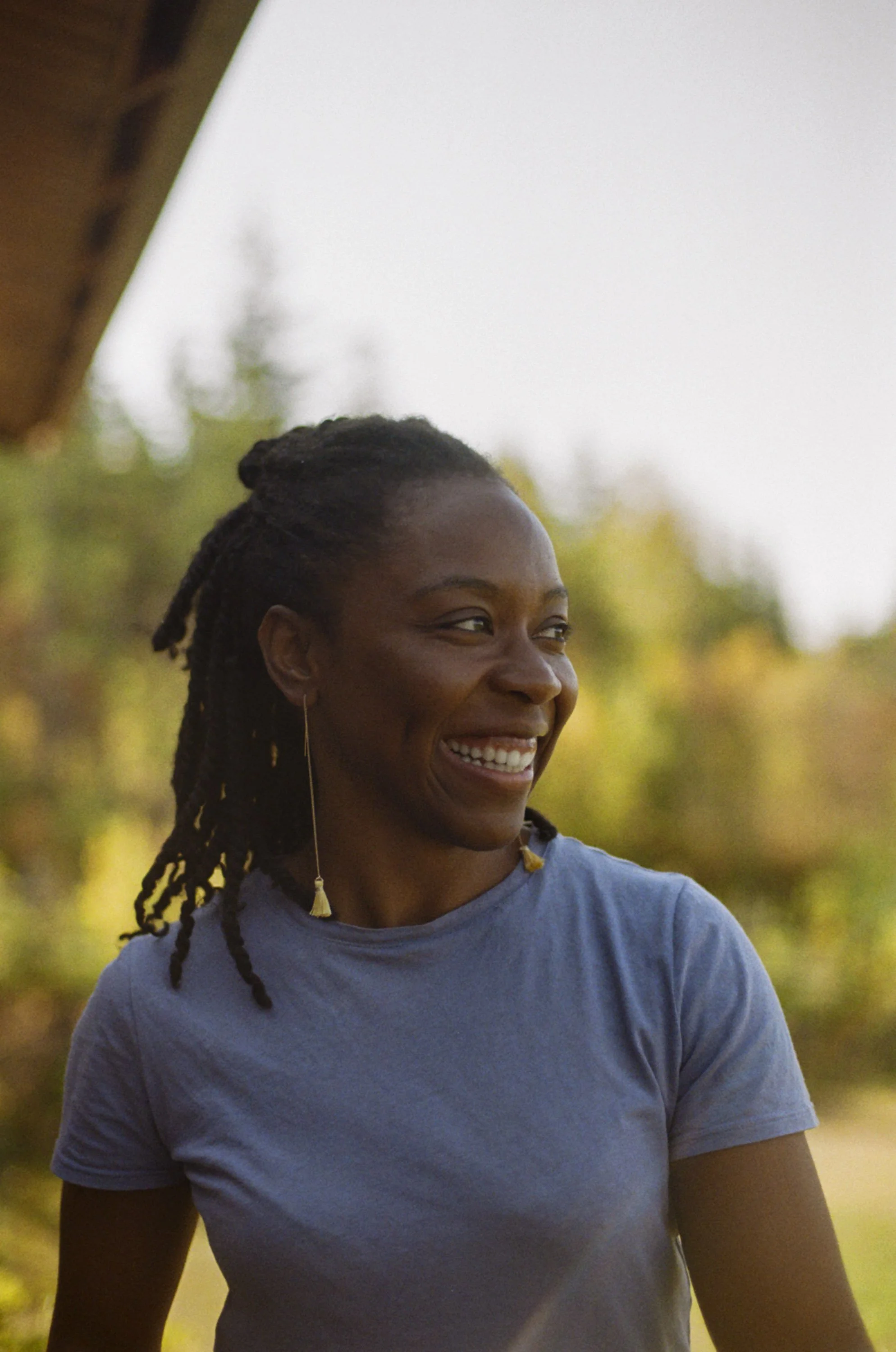 A woman with braided hair smiling outdoors during daytime, wearing a light purple shirt, with blurred trees and sunlight in the background.