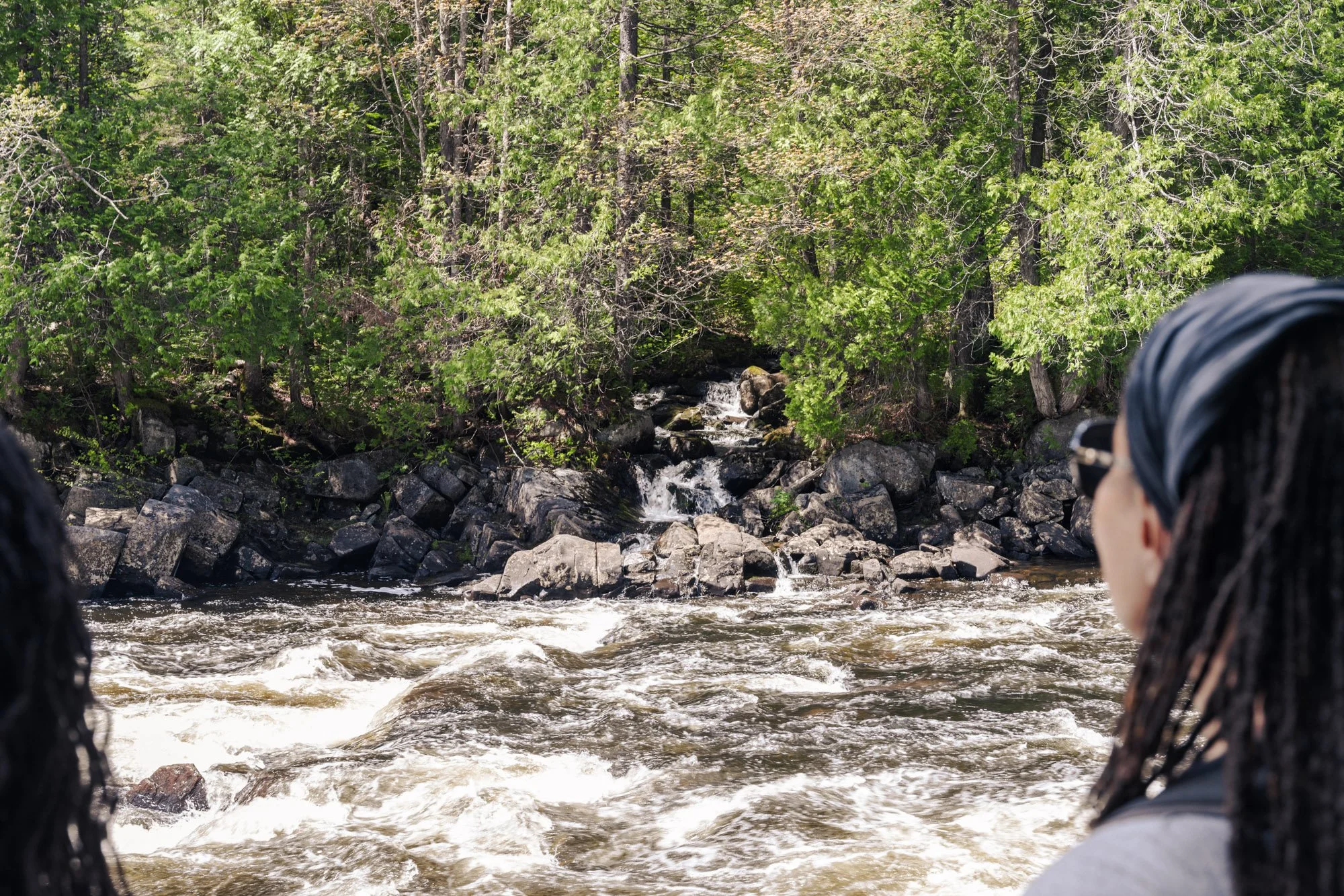 Two people wearing dark clothing and head coverings sitting near a river, surrounded by green trees and rocks.