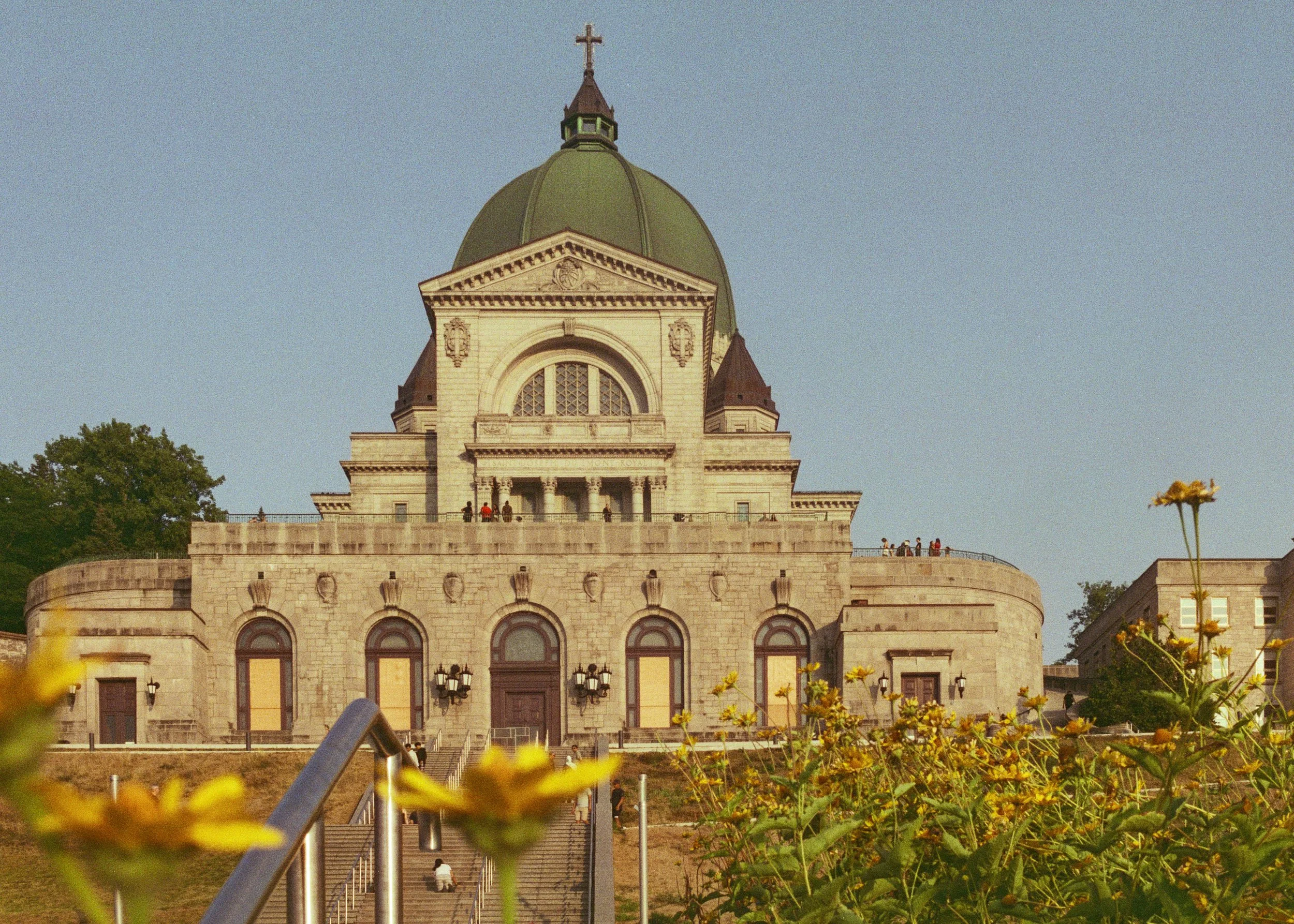 The image shows a large historic building with a prominent green dome topped with a cross. There are stairs and railings leading up to the building's entrance, and several people are walking and standing around the area. The foreground includes some 