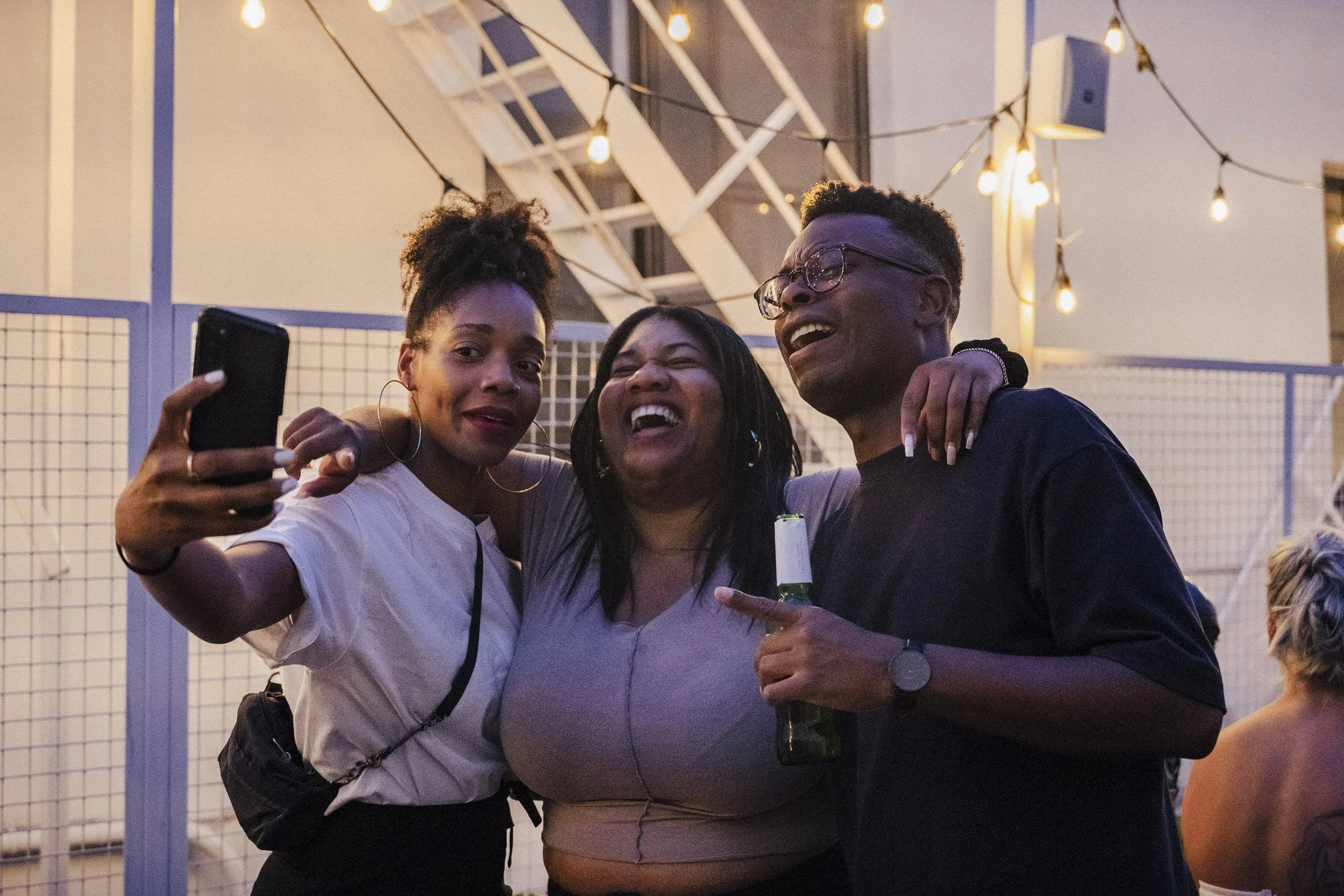 Three women are taking a selfie together at an outdoor event during the evening, with string lights overhead and a white metal fence in the background. They are smiling and enjoying each other's company, holding drinks, and posing closely.