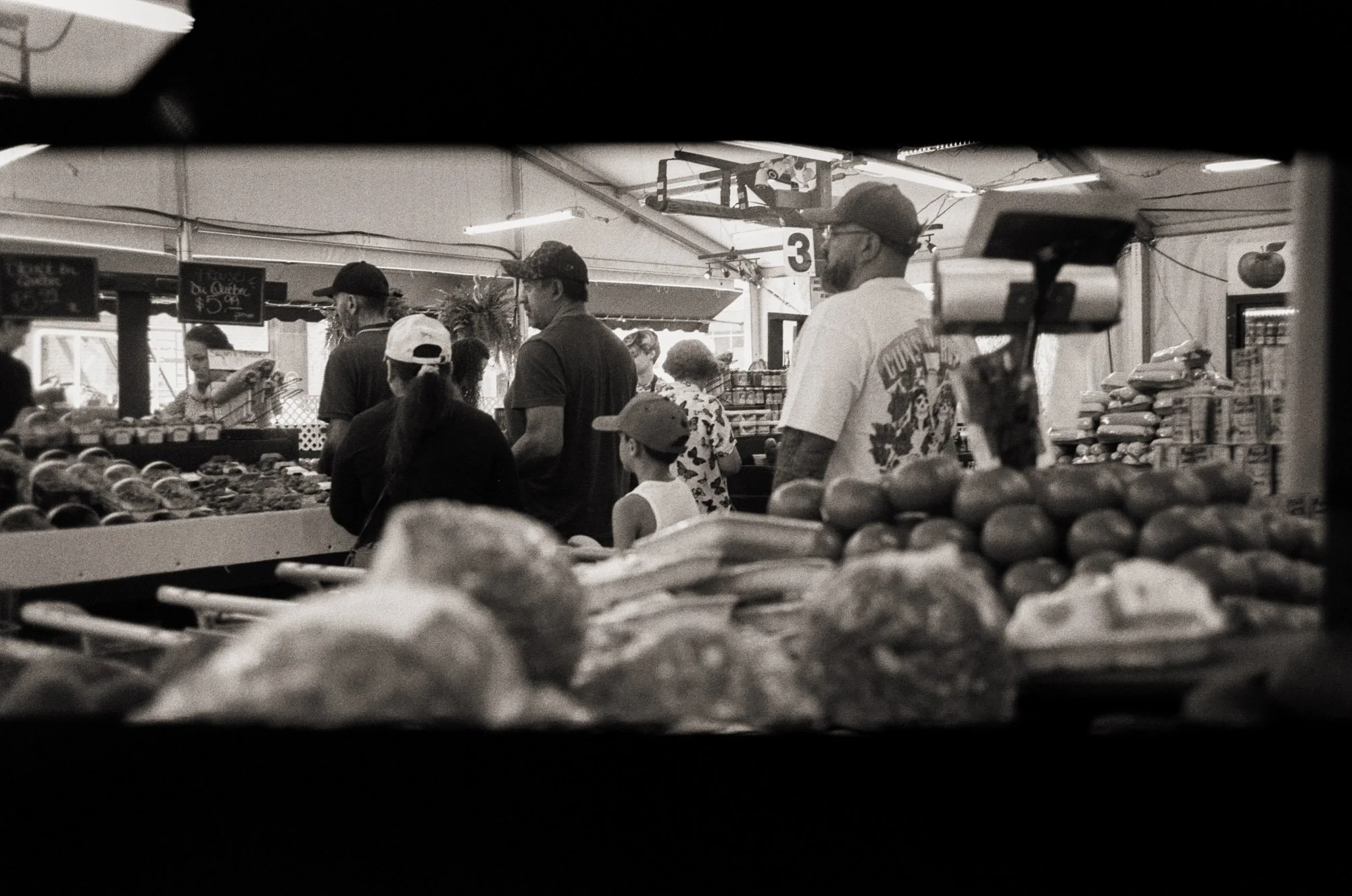 People shopping at an indoor market, focusing on the produce section with various fruits and vegetables. The scene includes customers and vendors, with signs indicating prices, in black and white.