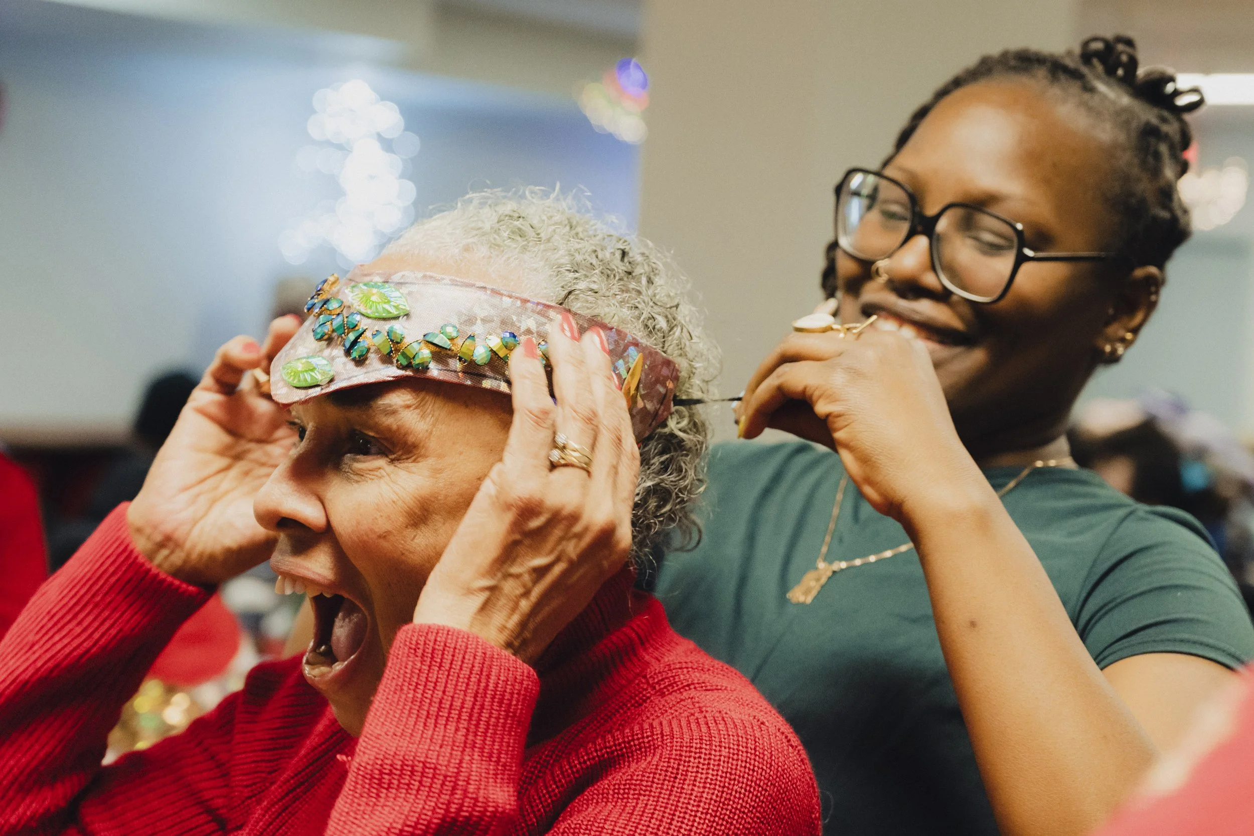 An elderly woman with curly gray hair is smiling and holding her head with her hands, wearing a colorful headband with large decorative elements, while a young woman with braided hair and glasses is helping her, possibly with a beauty or health treat