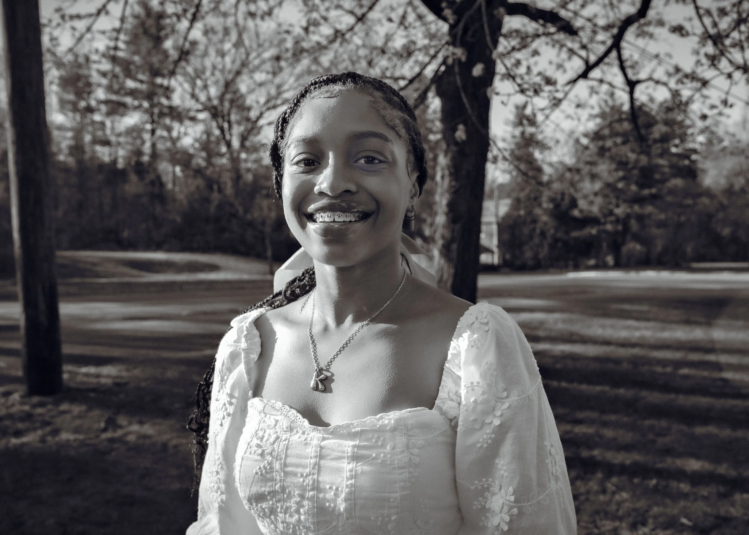 A young woman smiling outdoors in a park with trees in the background, wearing a white dress with floral embroidery and a necklace.