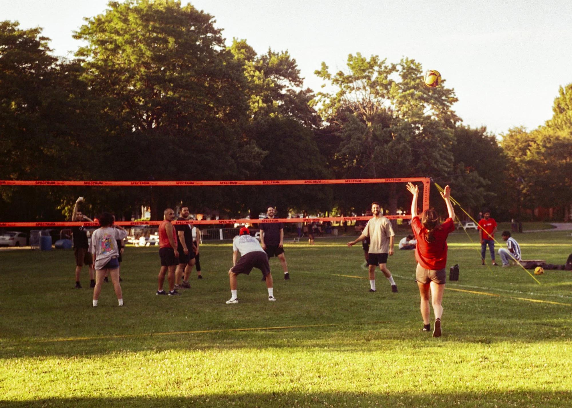 People playing volleyball outdoors on a grassy field during sunset, with trees in the background.